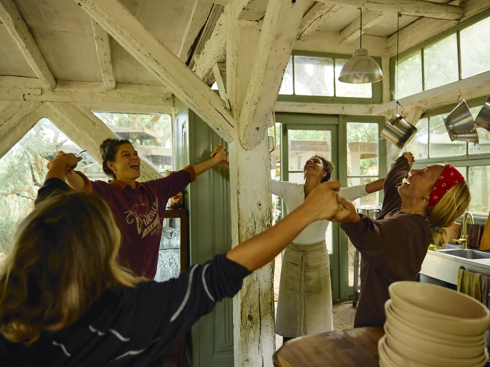 Grupo de mulheres celebrando e dançando juntas em uma cozinha iluminada naturalmente, com janelas grandes e utensílios de cozinha pendurados no teto.