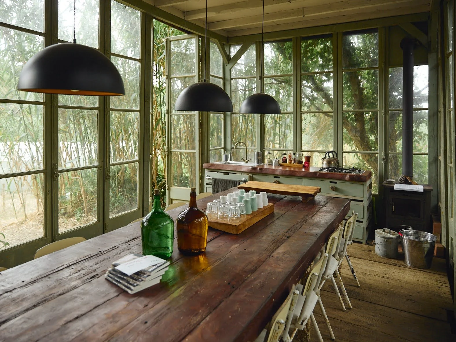 Cozinha rústica com mesa de madeira, cadeiras brancas, luminárias pretas suspensas, janelas grandes de vidro, pia, utensílios de cozinha, garrafas de vidro coloridas, copos e livros sobre a mesa, vista para vegetação ao ar livre.