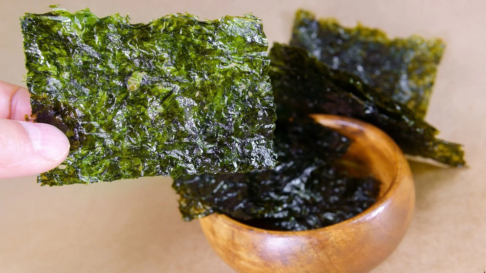 Folha de alga marinha seca sendo segurada por uma mão, com um recipiente de madeira ao fundo.
