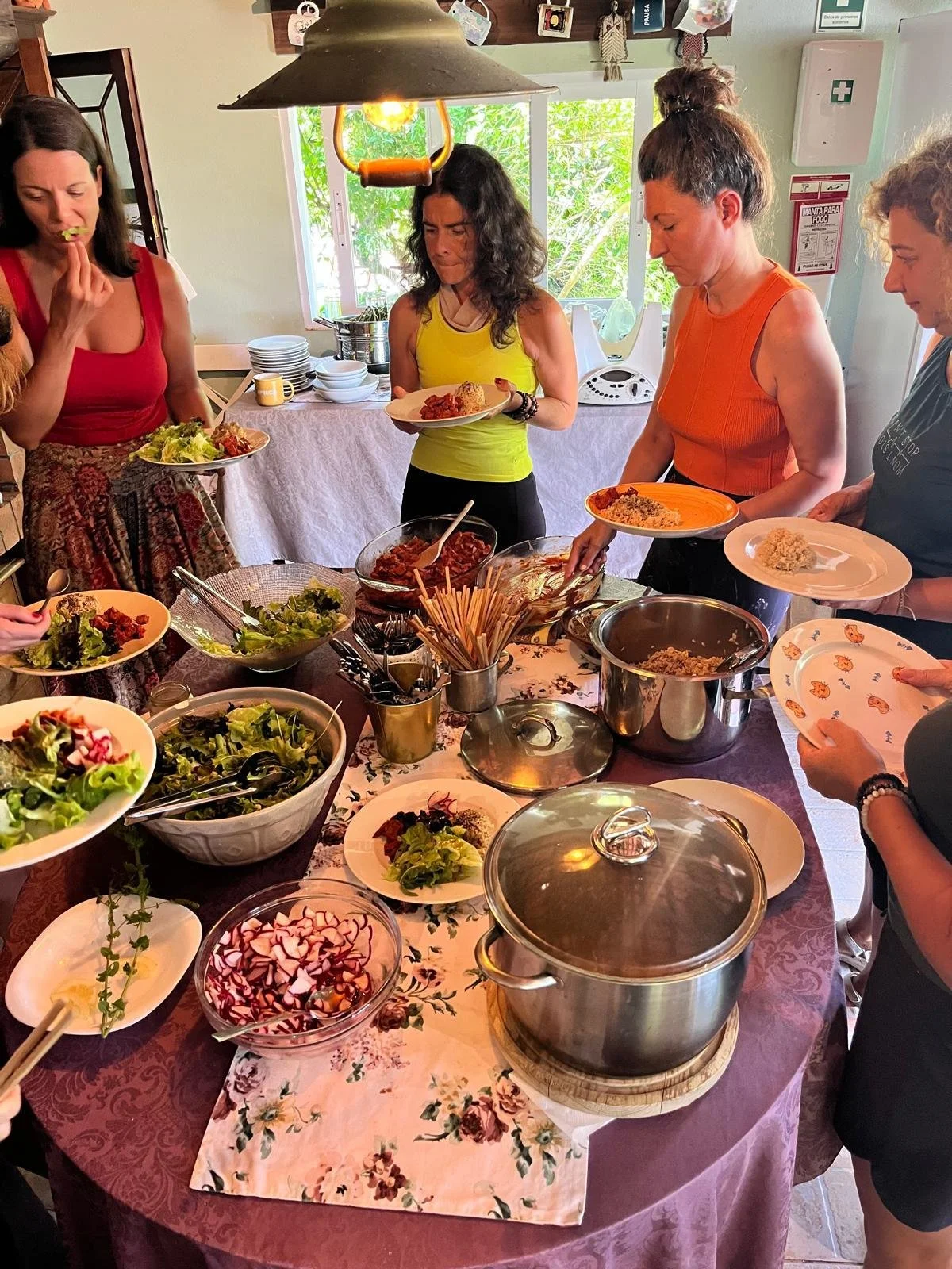 Mulheres servindo-se em uma mesa de comida com saladas, pratos quentes, cubos de radish, tigelas de utensílios e uma panela com tampa em um ambiente bem iluminado com janelas e decoração de parede.