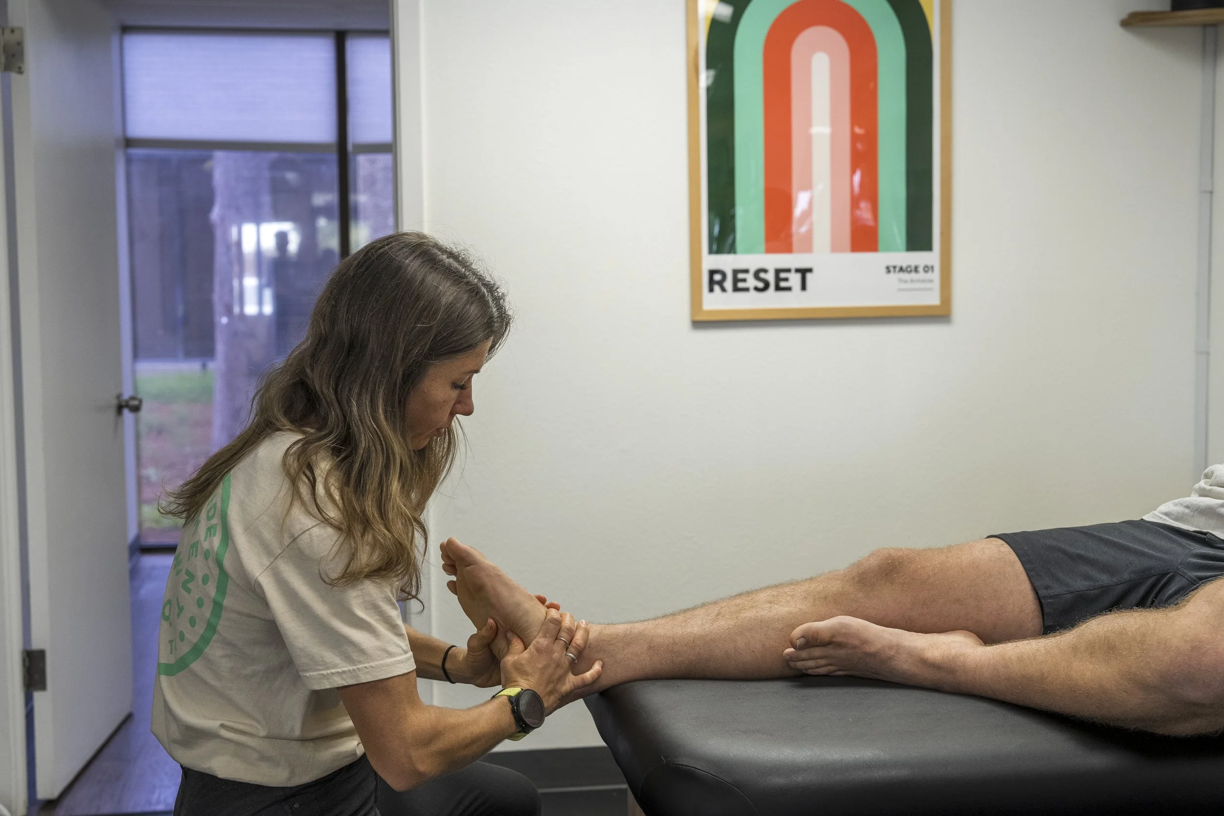 A woman is performing a medical procedure on a man's arm, possibly taking blood or administering an injection, in a clinical setting with a colorful 'RESET' poster on the wall.