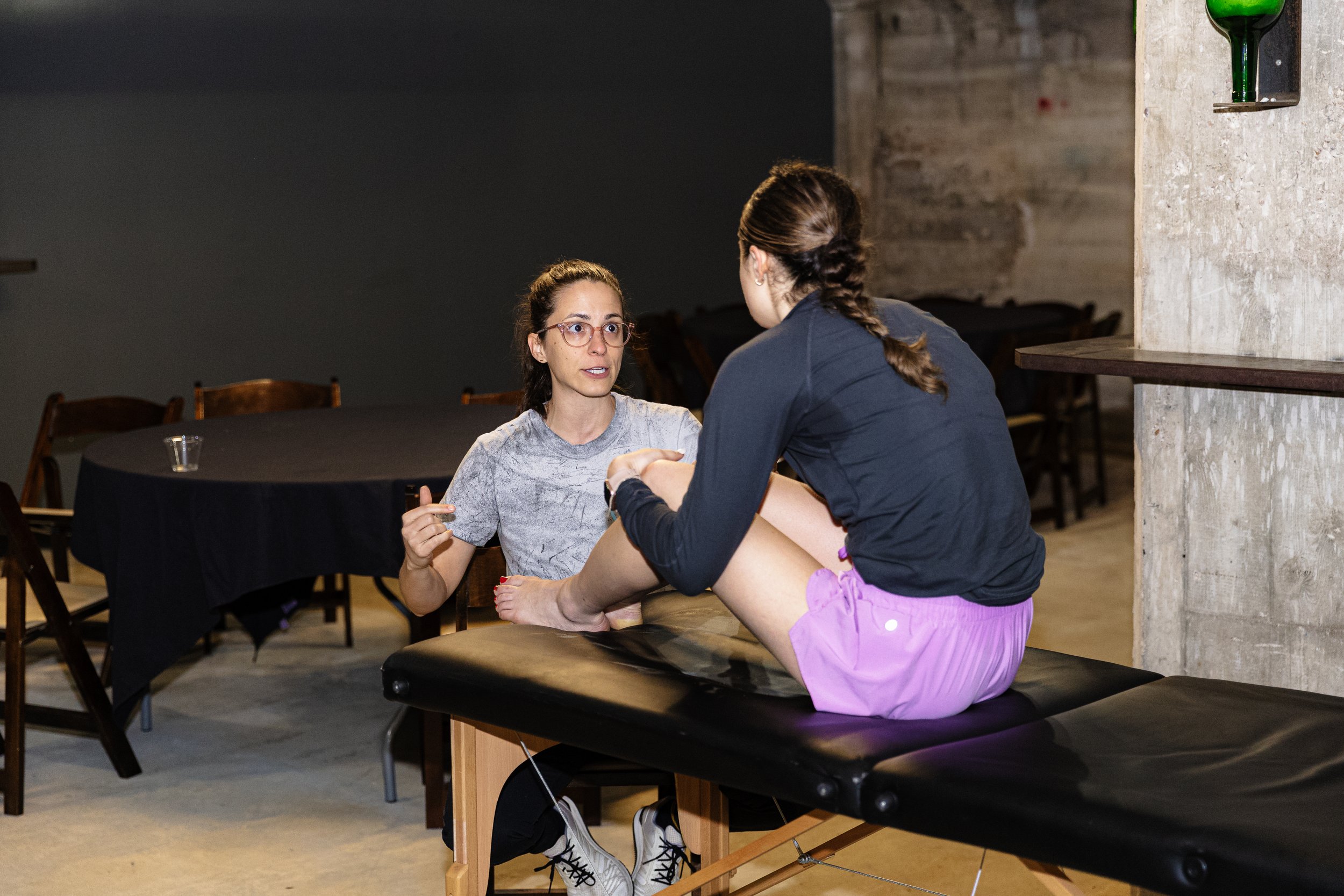 A woman with glasses and a gray T-shirt sits at a table, talking to a girl who is balancing on a black massage table with her legs crossed and wearing pink shorts and a dark long-sleeve shirt.