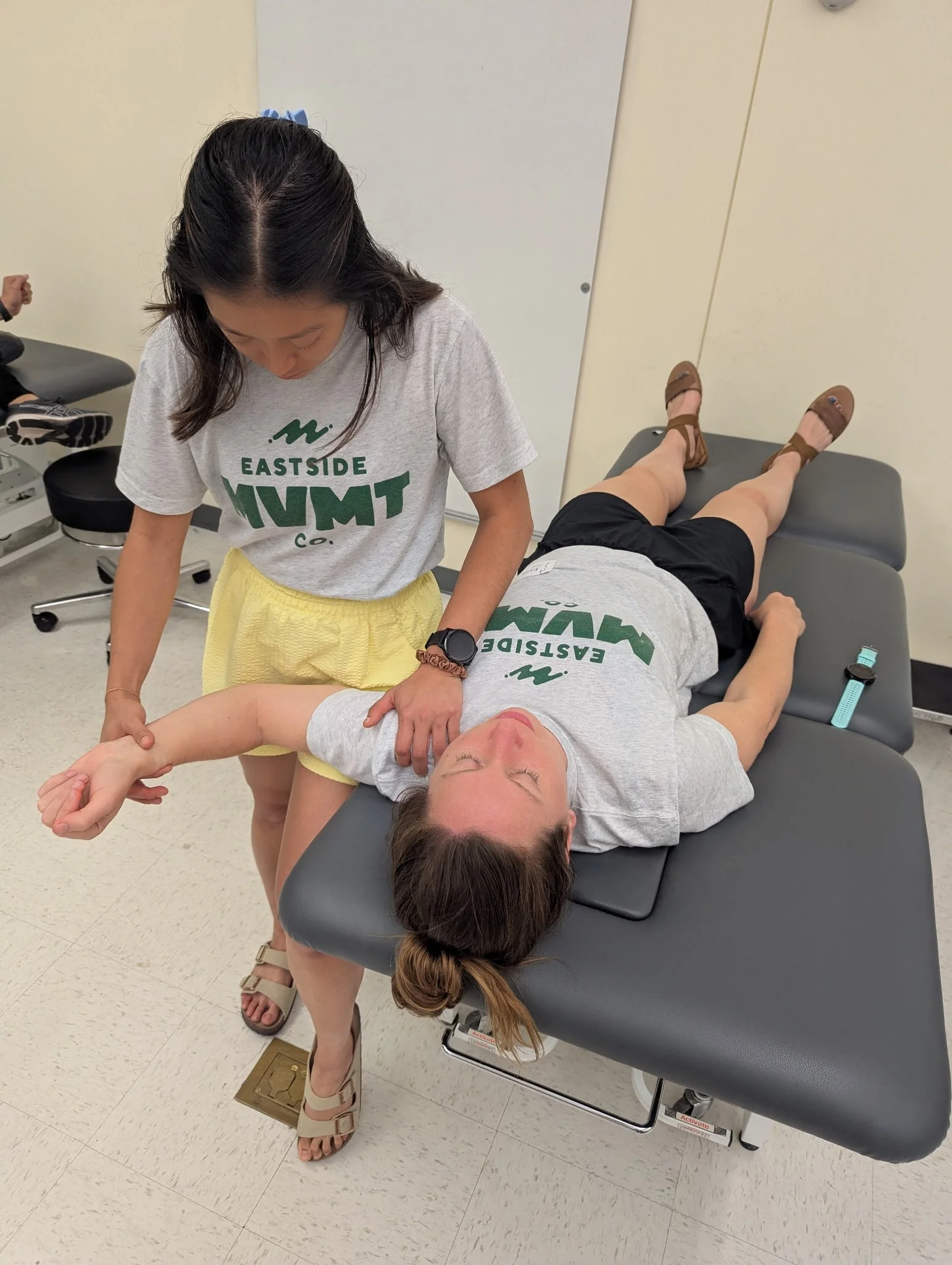 Woman lying on a medical examination table with healthcare provider standing beside her, holding her arm, in a clinical setting.