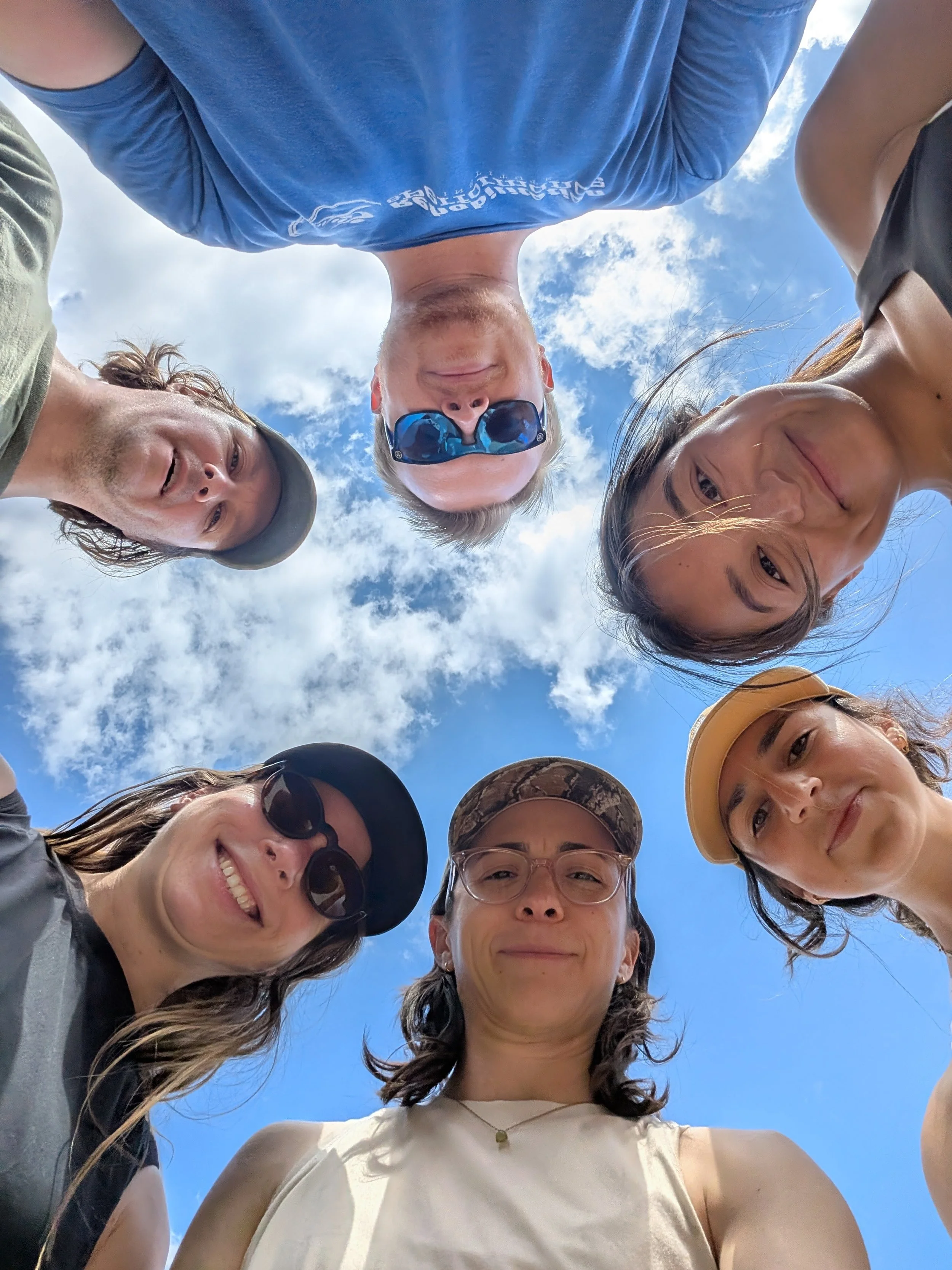 Group of people in a circle looking down at the camera, taken from below with a bright blue sky and clouds in the background.