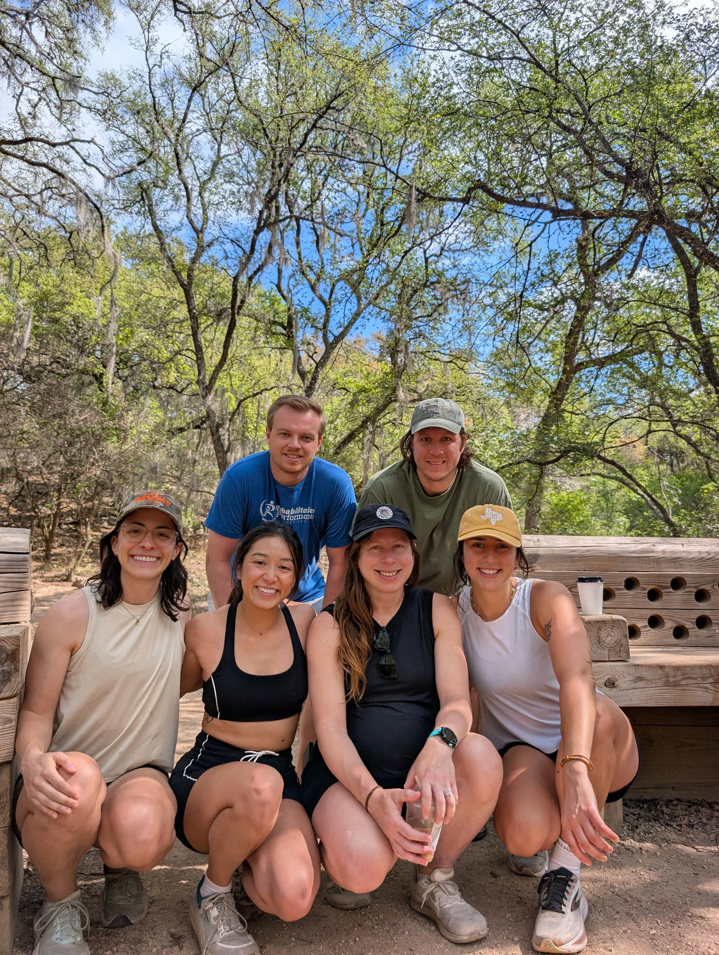 A group of eight young adults smiling and posing outdoors, with trees and a bright blue sky in the background.