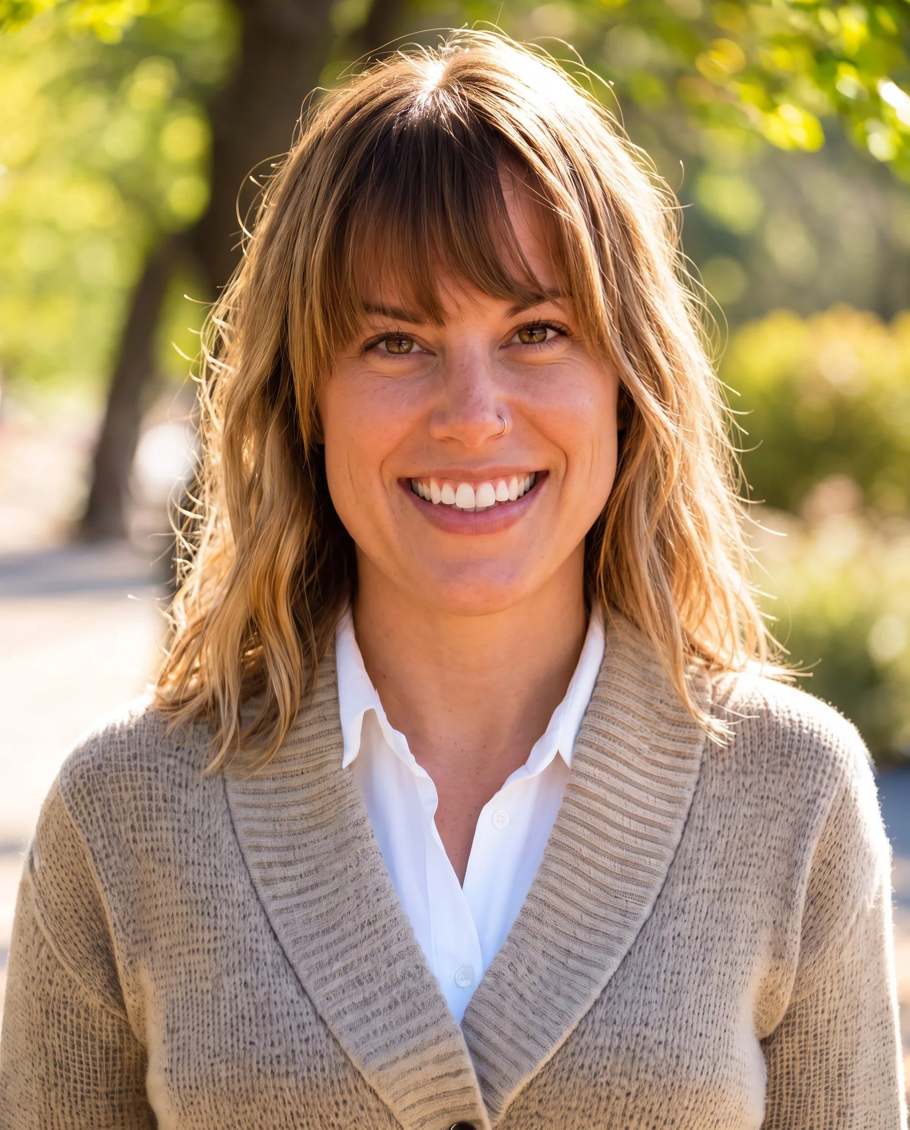 A woman smiling outdoors with sunlight and green trees in the background. She has brown hair, a pierce in her nose, and is wearing a white shirt with a beige knit cardigan.