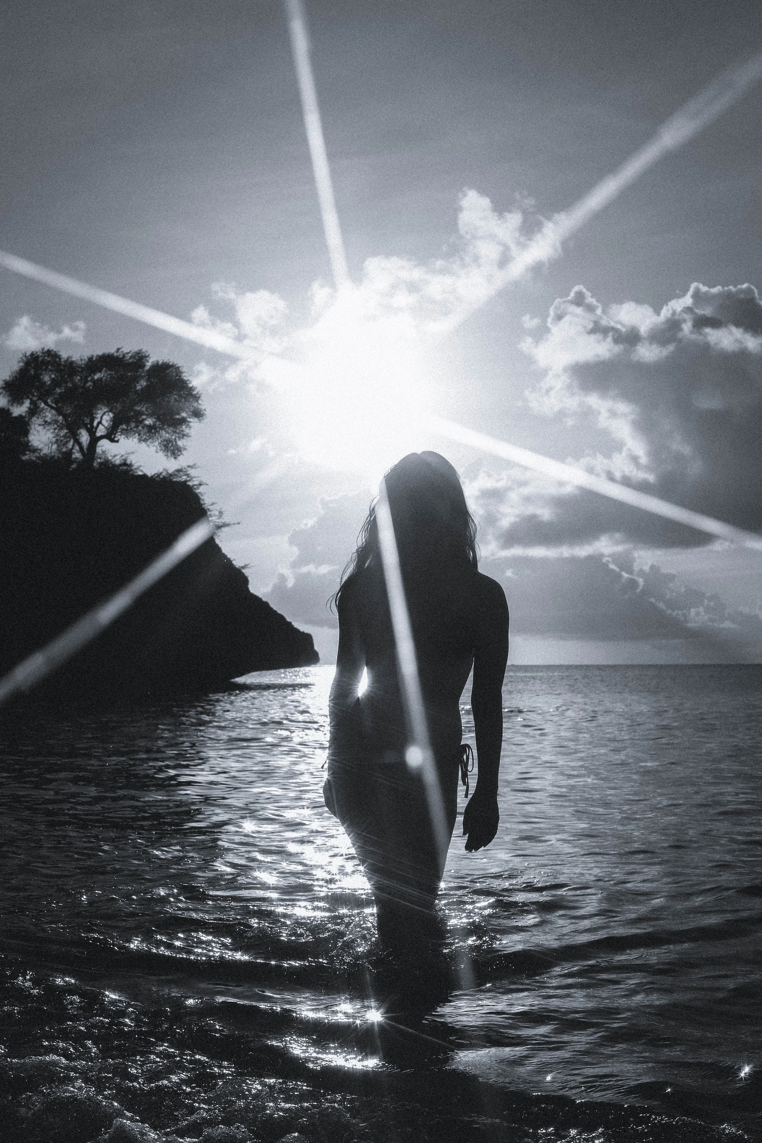A silhouette of a woman wading in the water on the beach with the sun shining behind her, creating a dramatic black-and-white scene.