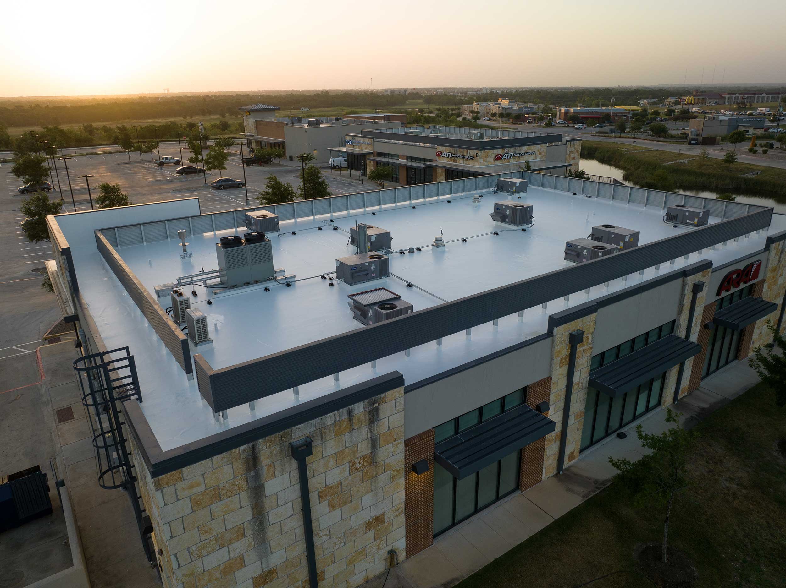 Aerial view of a commercial building with a flat white roof dotted with HVAC units, surrounded by parking lots, trees, and other retail stores at sunset.