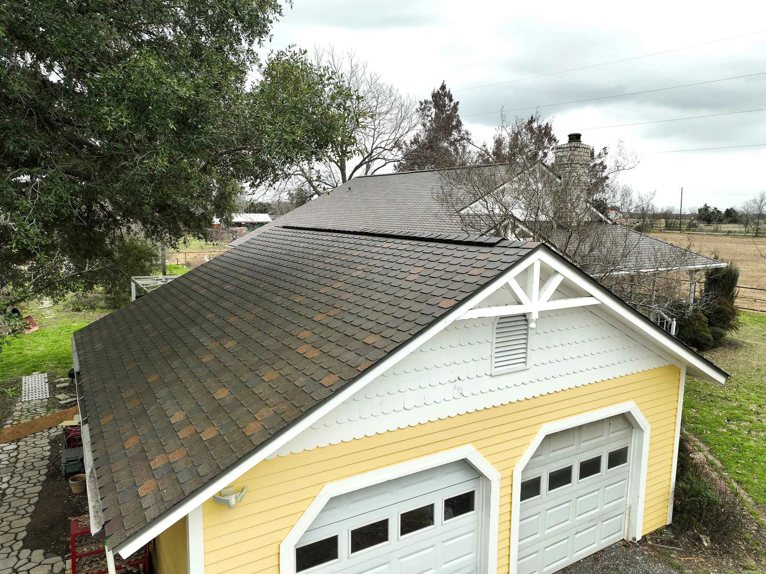 A yellow garage with white trim and two white garage doors, situated beneath a gable roof with gray shingles, with trees and a cloudy sky in the background.