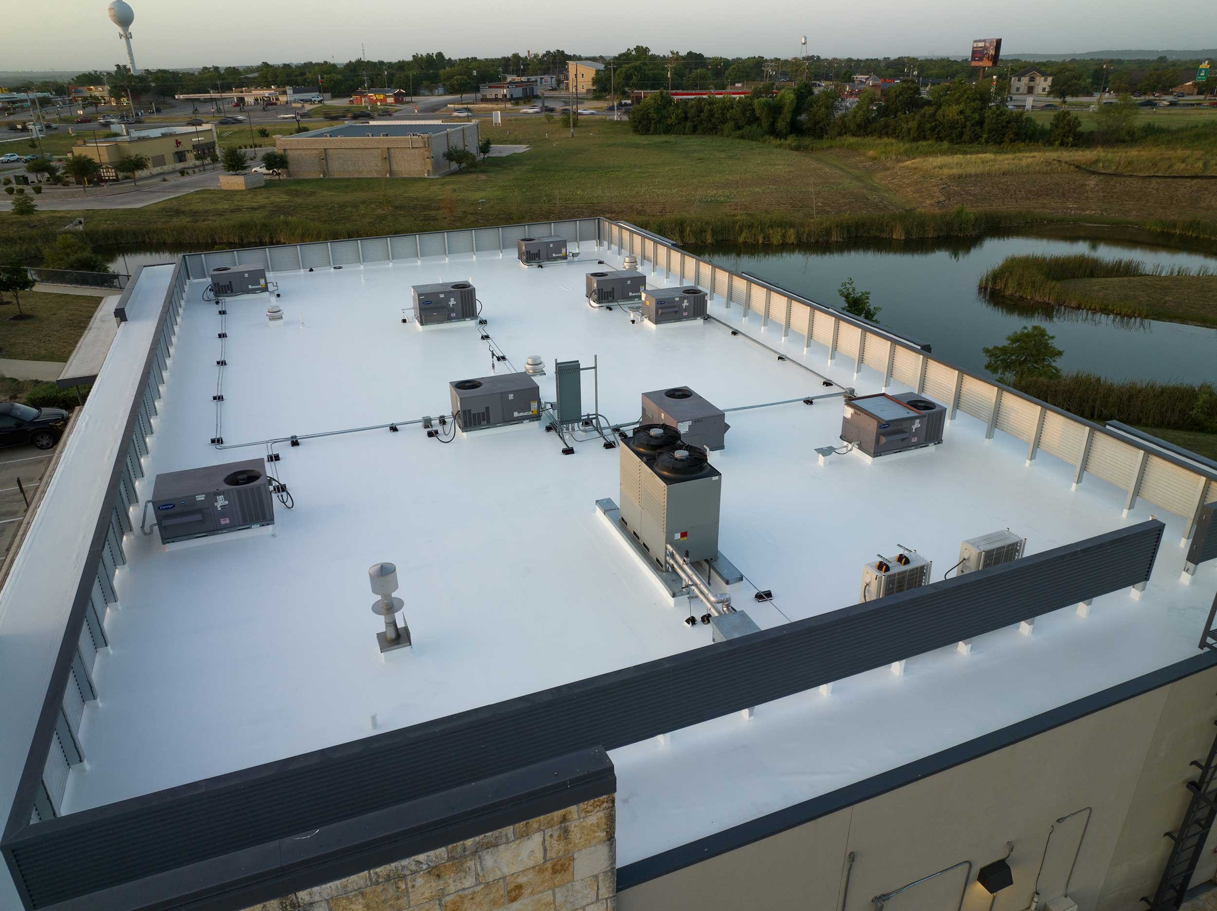 Rooftop view of a building's white flat roof featuring multiple HVAC units, a vent, and a small fan, with a background of trees, a pond, and a busy street with commercial buildings and billboards.