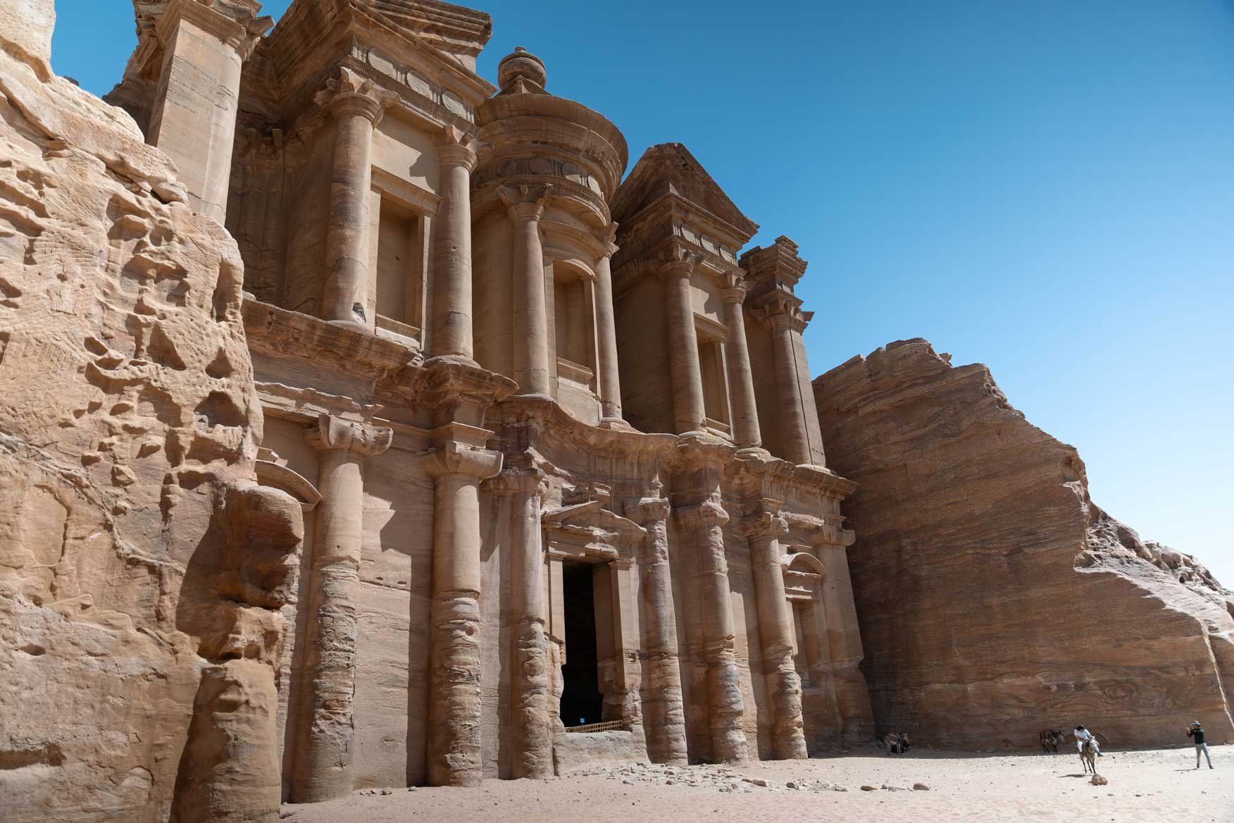 The famous Monastery of Petra in Jordan, carved into sandstone cliffs, with people and camels in the desert.