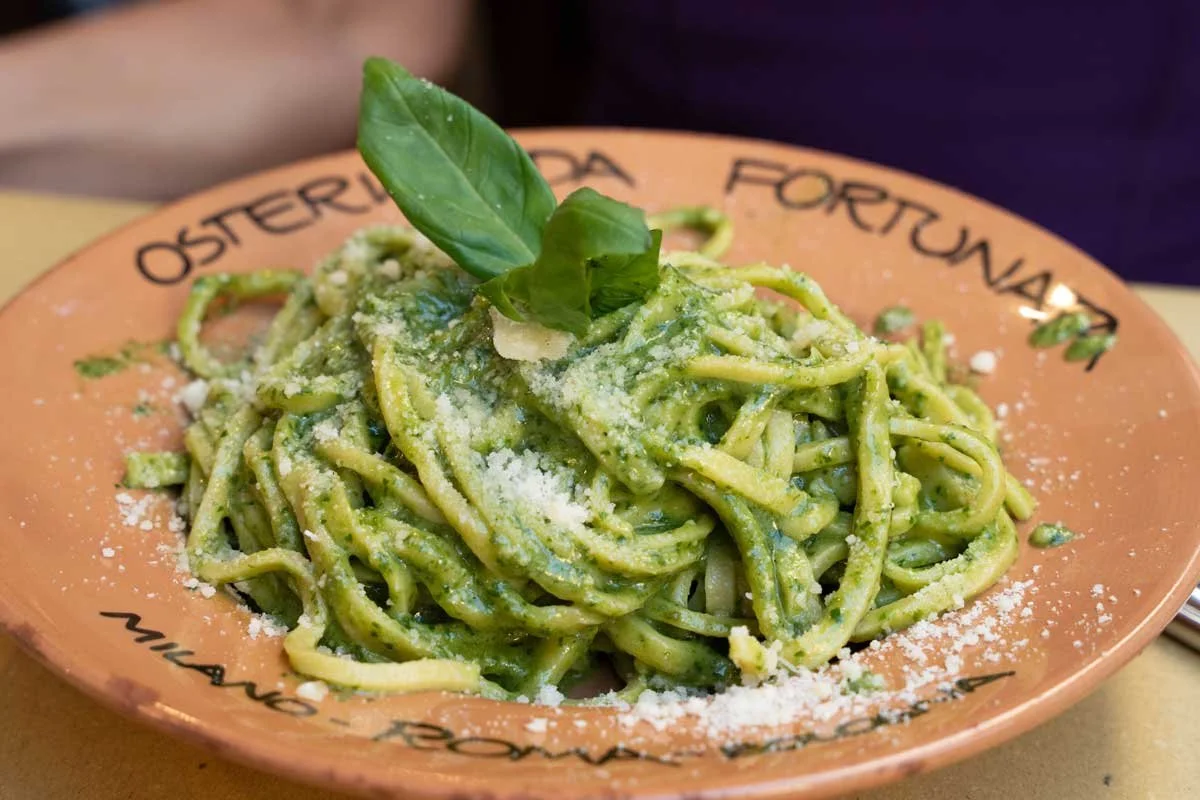 A plate of green pesto pasta garnished with a basil leaf and grated cheese, on a peach-colored plate with words written around the rim.