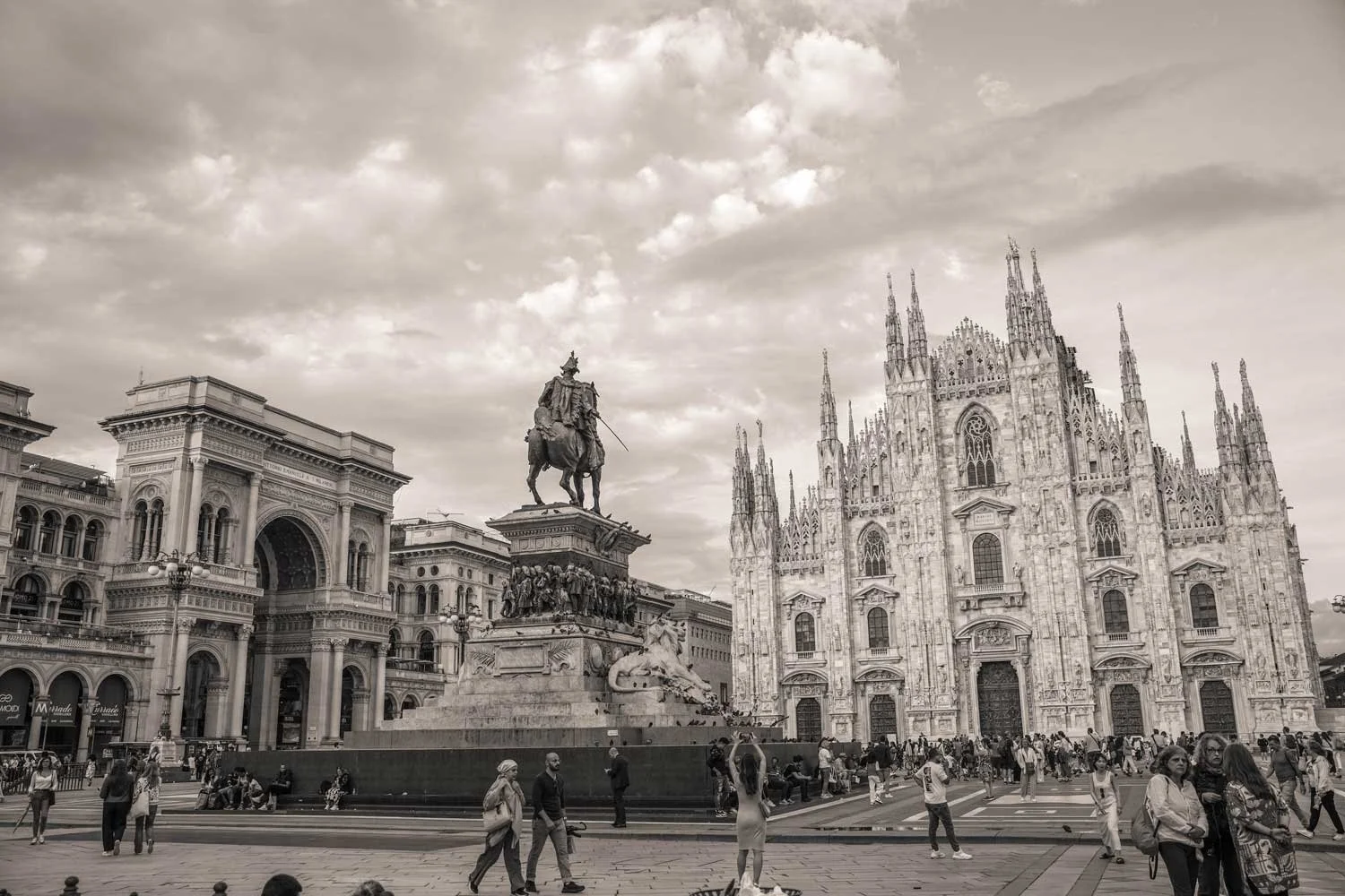A busy city square featuring a large equestrian statue, historic buildings, and the Gothic-style Milan Cathedral in the background, with people walking and gathering in the area.
