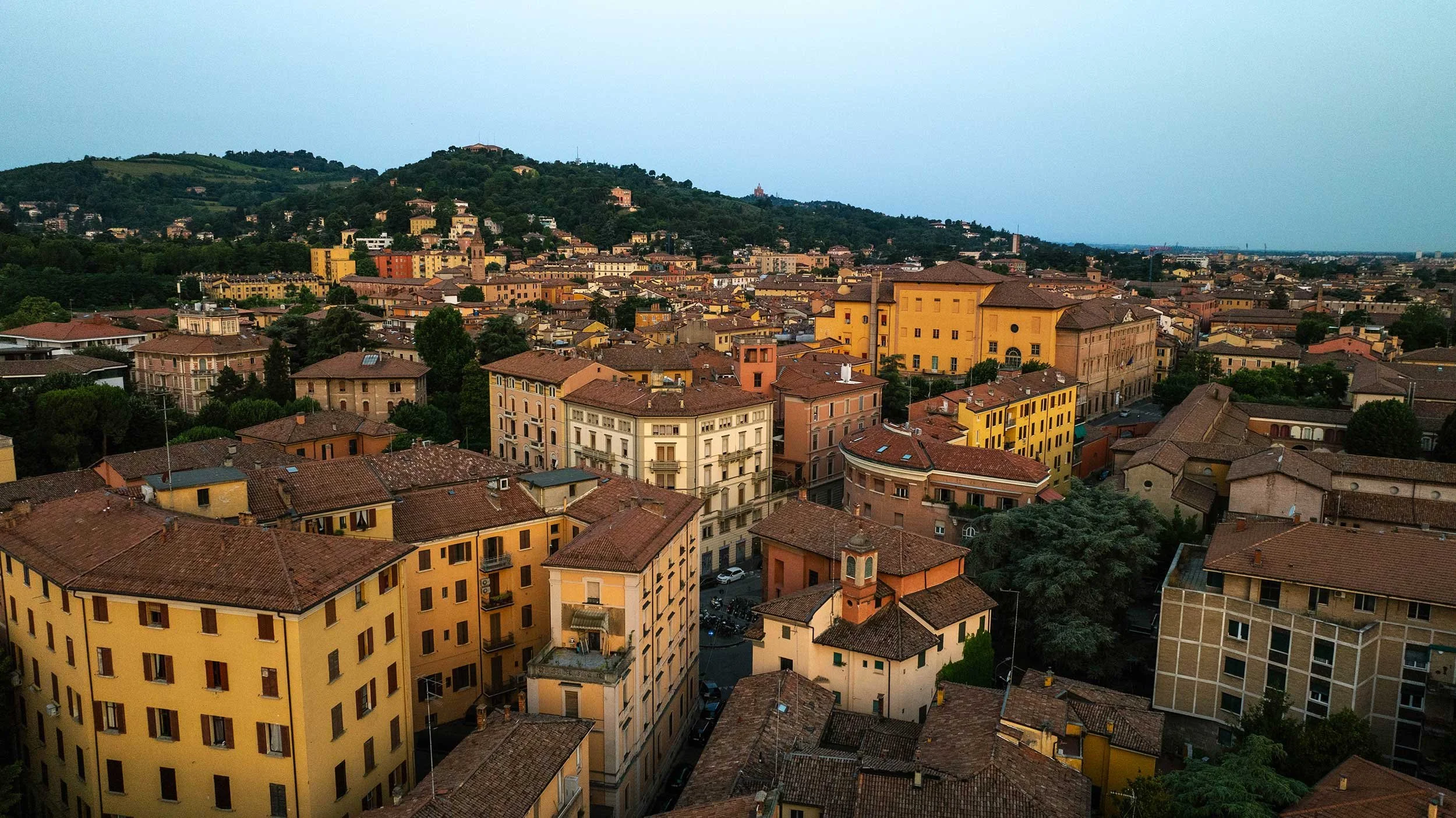 Aerial view of an Bologna with colorful buildings, red-tiled roofs, and a green hillside in the background during daytime.