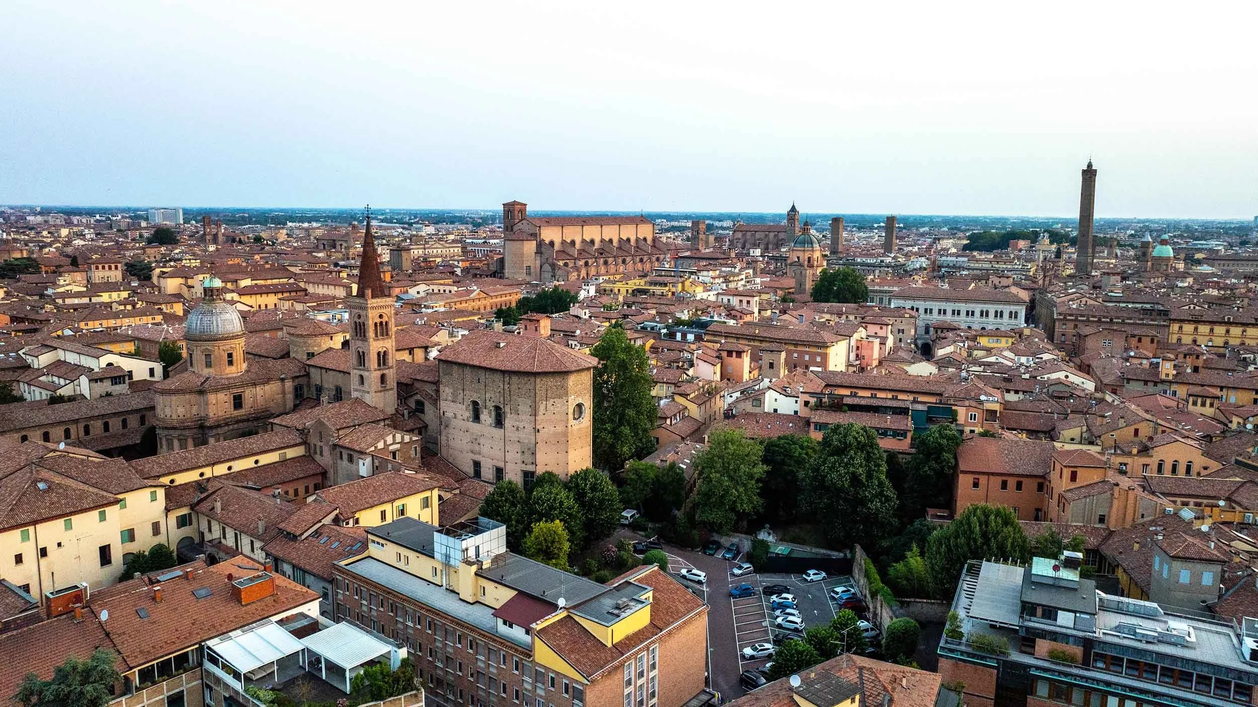 Aerial view of a historic Bologna with numerous buildings, church steeples, towers, and terracotta roofs, surrounded by green trees.