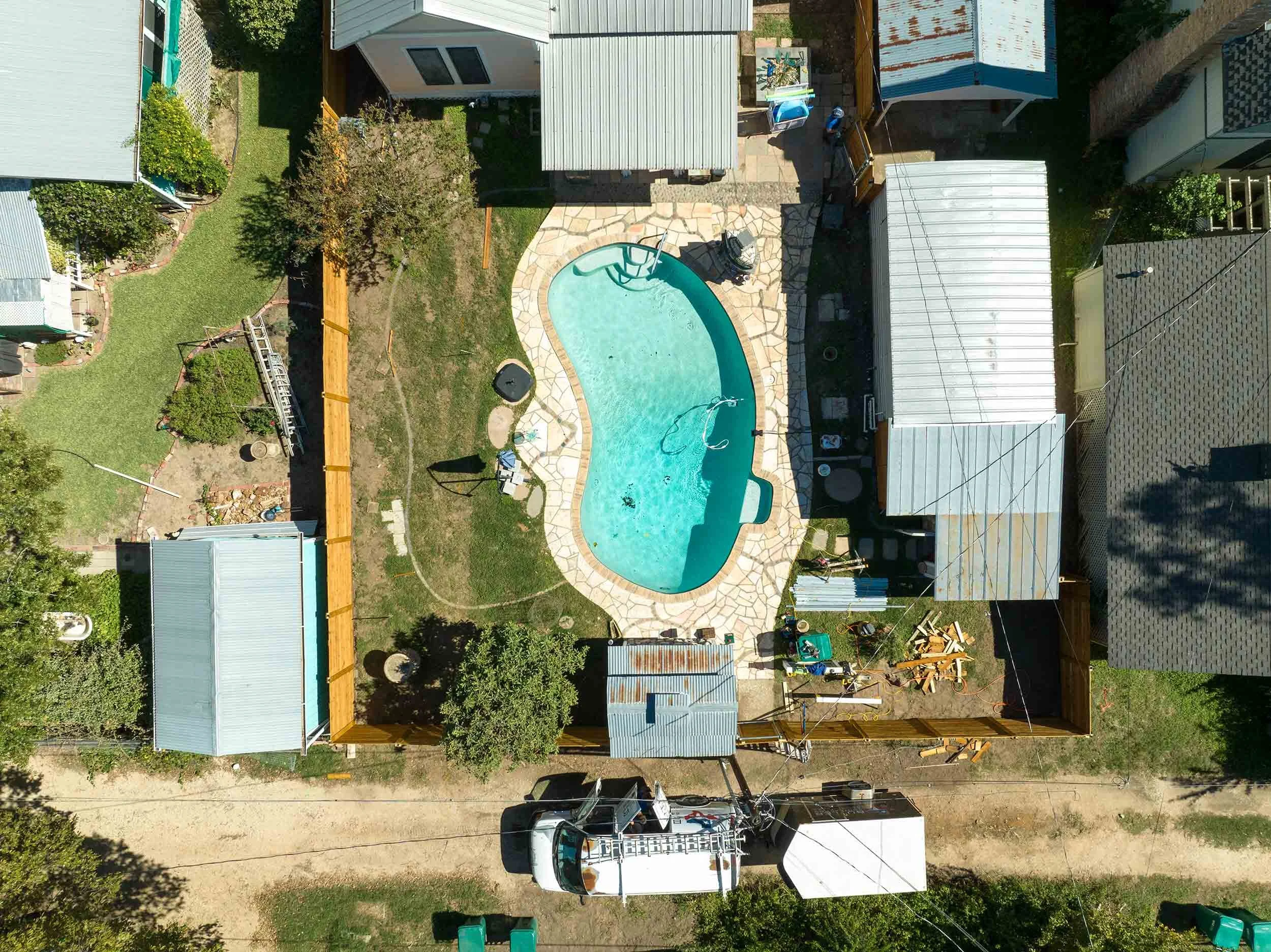 An aerial view of a backyard with a small swimming pool, surrounded by a stone deck, various sheds, and a lawn area with plants and trees. There are vehicles parked along the street at the bottom of the image.