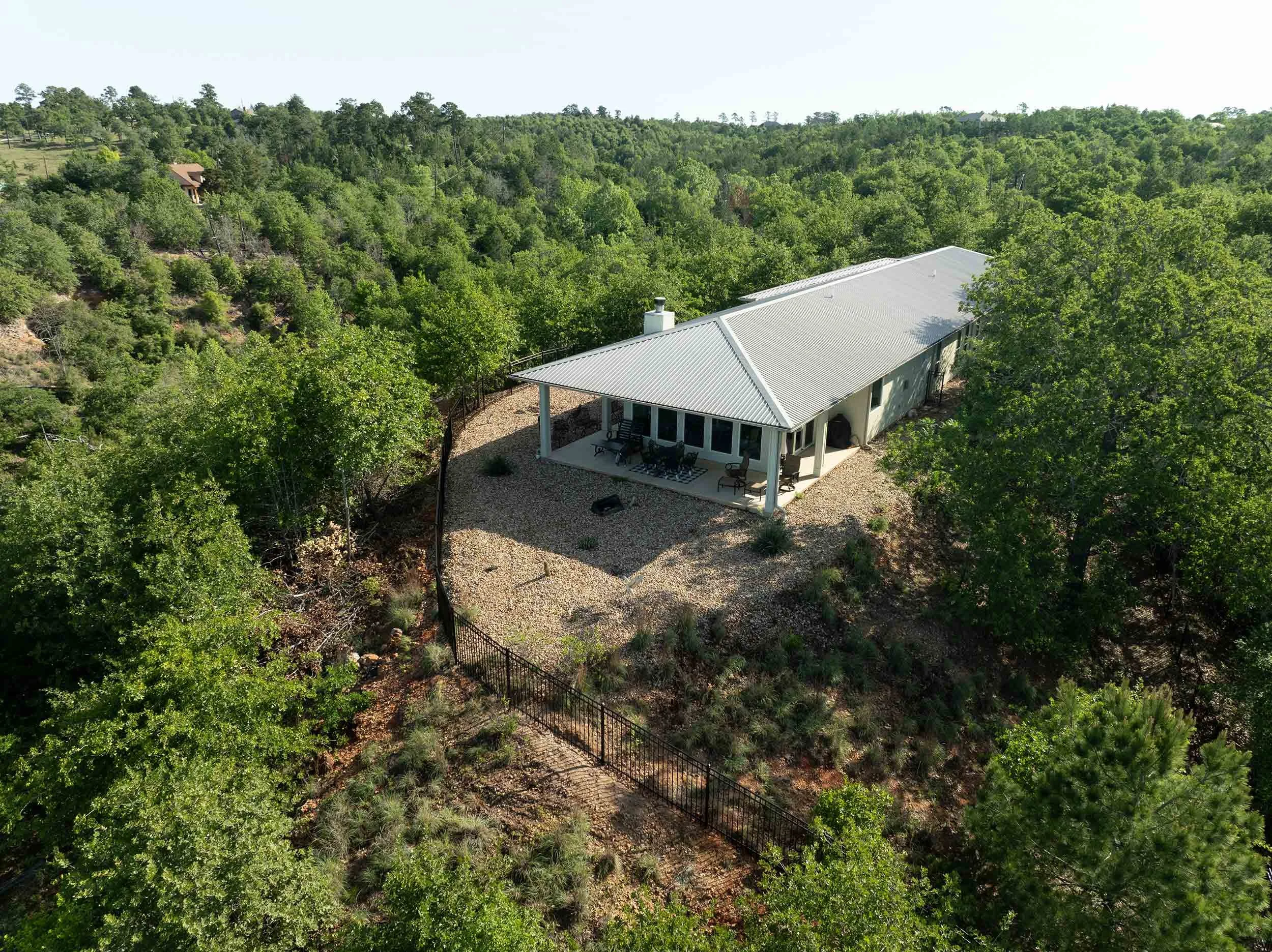 A house with a metal roof surrounded by lush green trees in a hilly forest landscape.