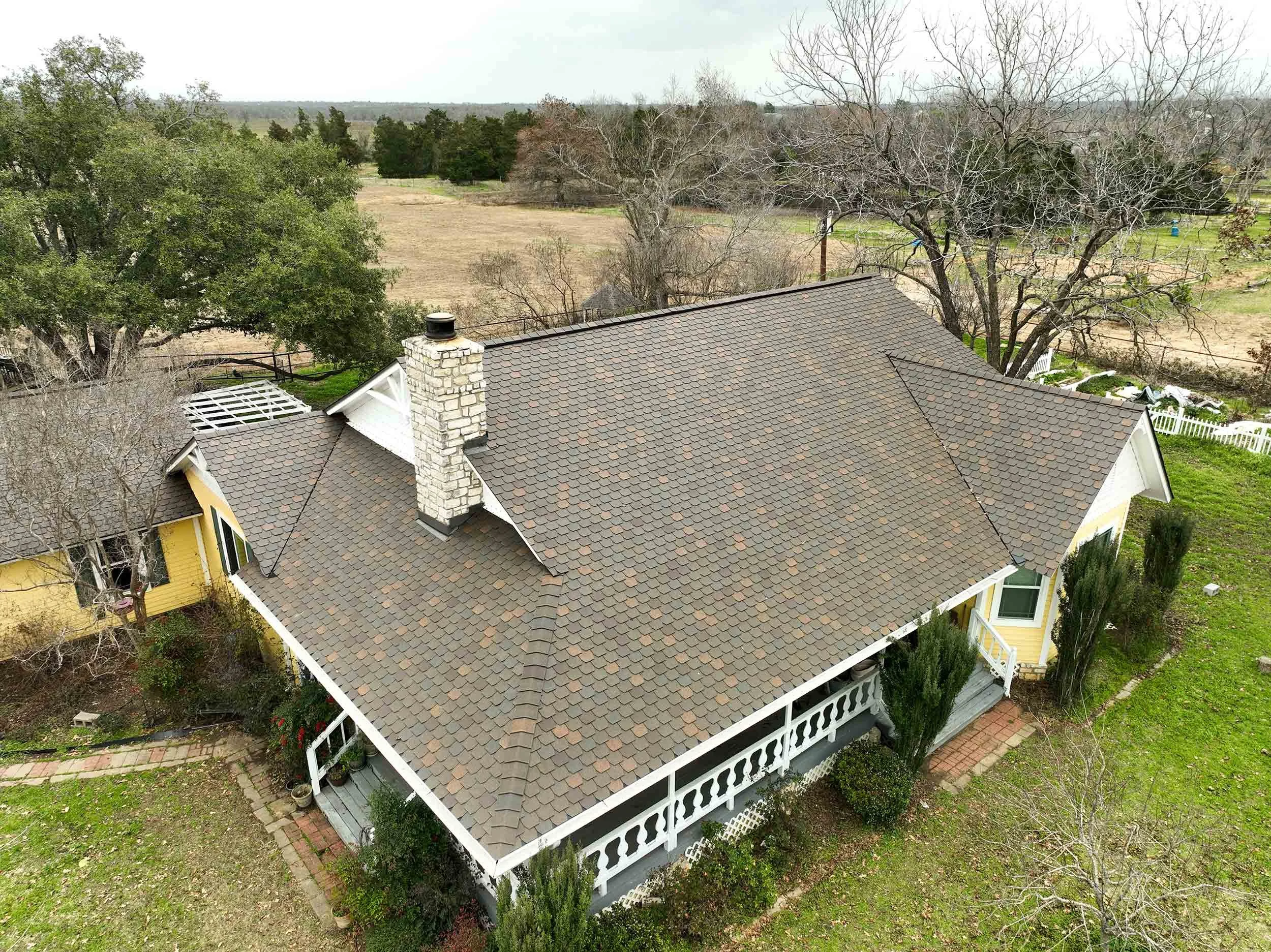 A yellow house with white trim and a porch, surrounded by trees and greenery, with a large shingled roof and a chimney. The house is set in a rural landscape with open fields and distant trees.