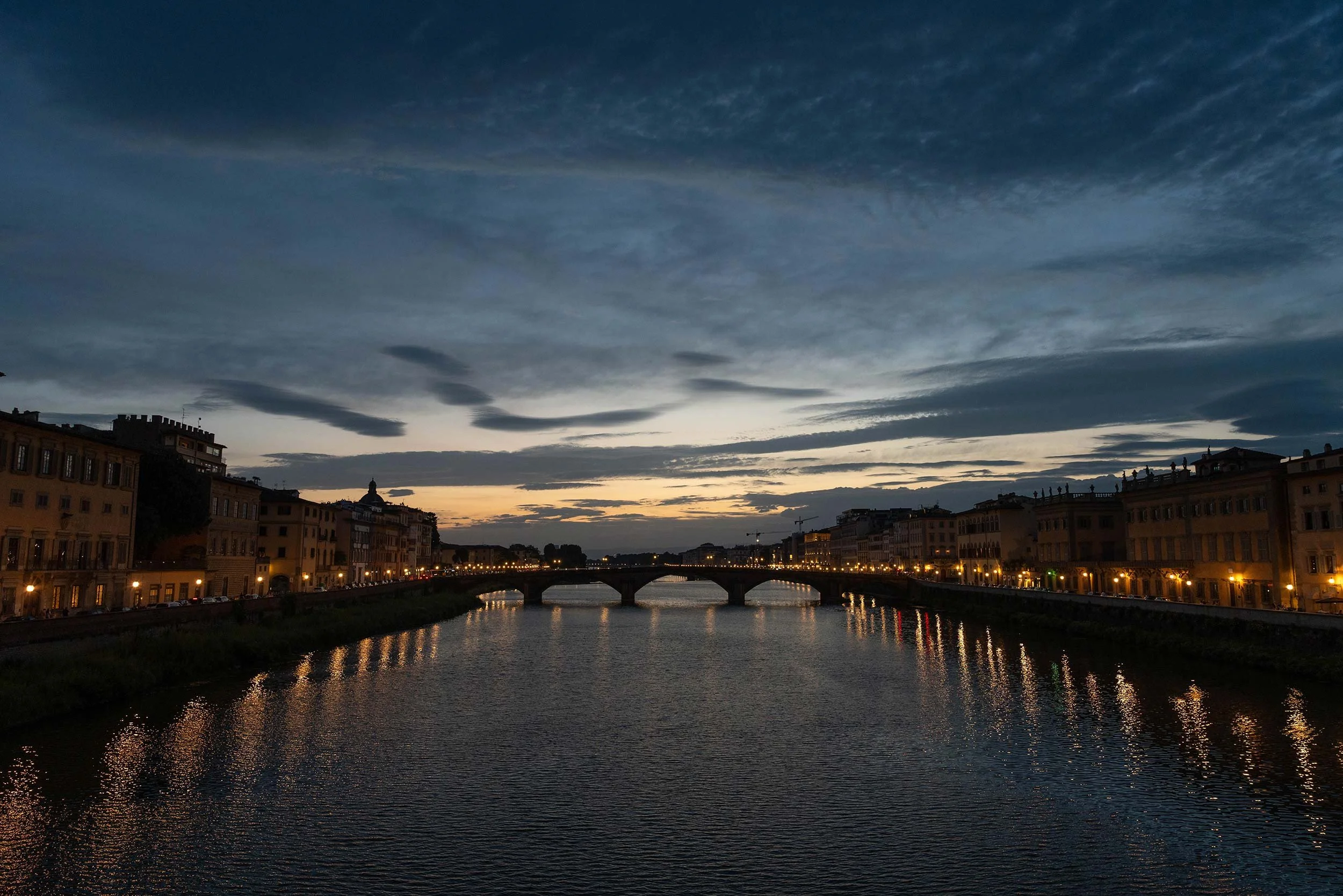 Florence, Italy cityscape at dusk over a river with illuminated buildings and a bridge, casting reflections on the water.