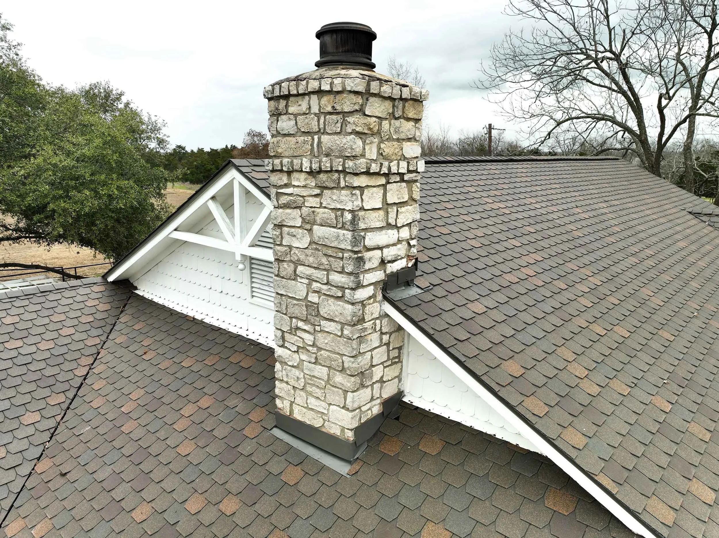 A house roof with brown shingles, a large stone chimney, and decorative white trim beneath the gable, with trees and a cloudy sky in the background.