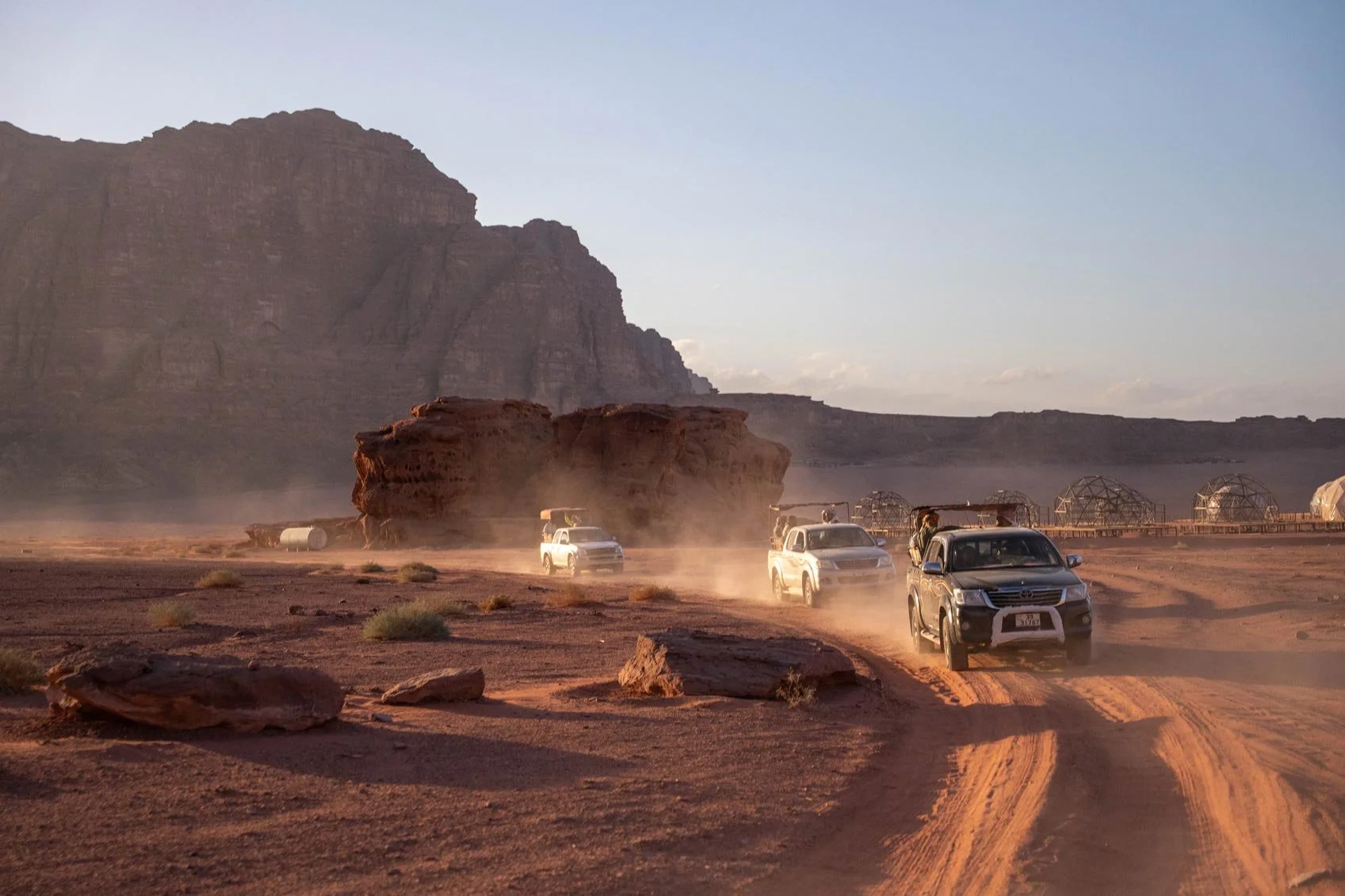 Four vehicles driving on a dusty desert road with rock formations and domed greenhouses in the background, under a clear sky.