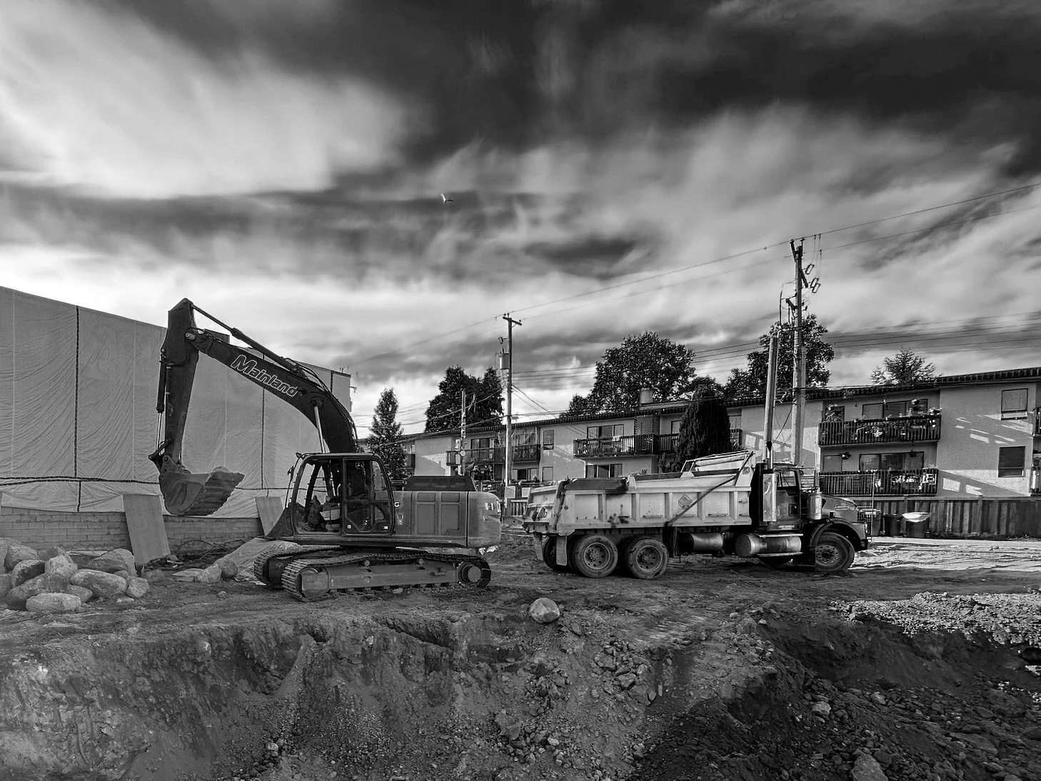 Excavator and dump truck performing earthworks during urban infill excavation and site preparation beside apartments