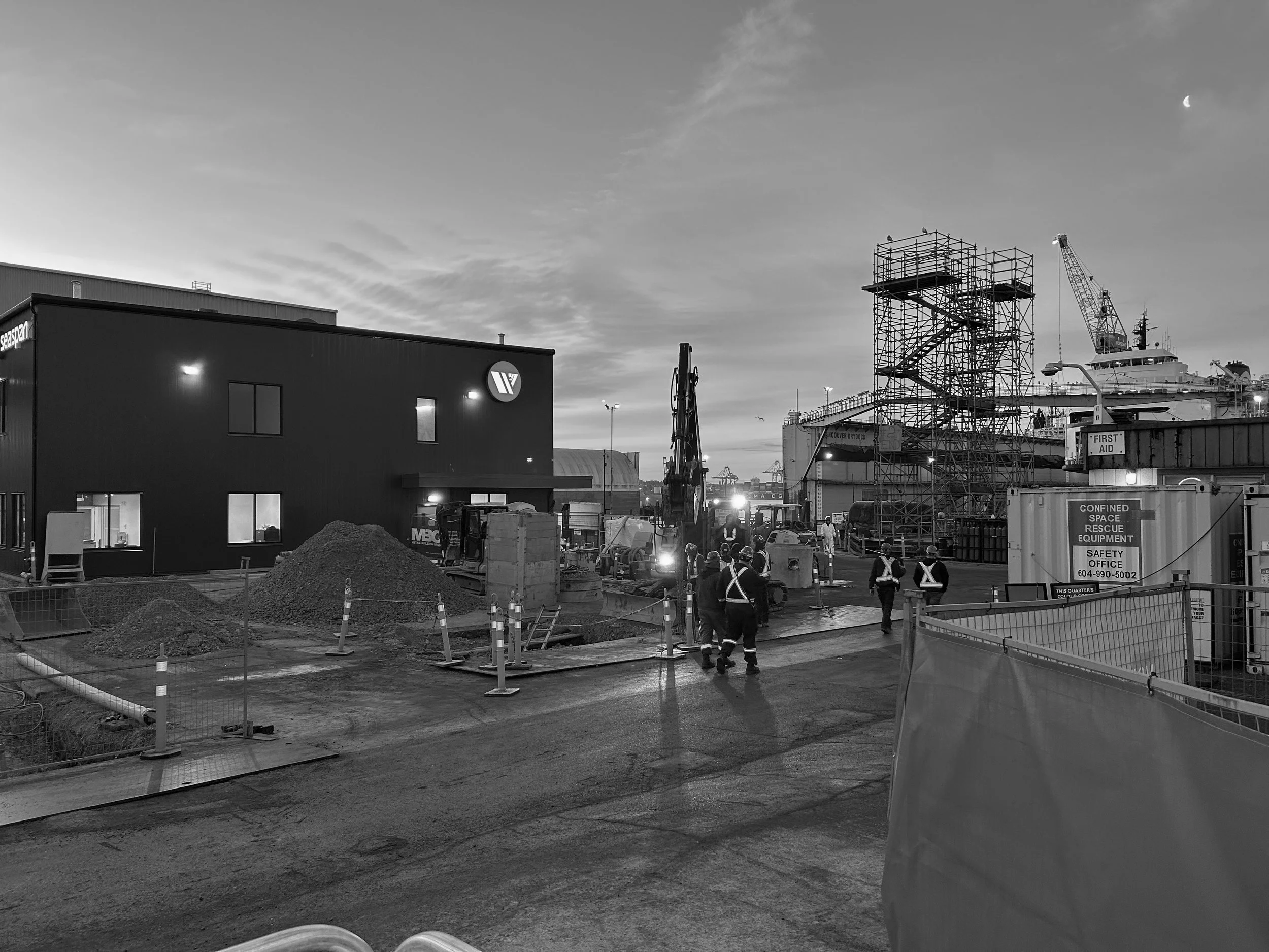 Crew working at a waterfront industrial site with scaffolding and excavator at dusk
