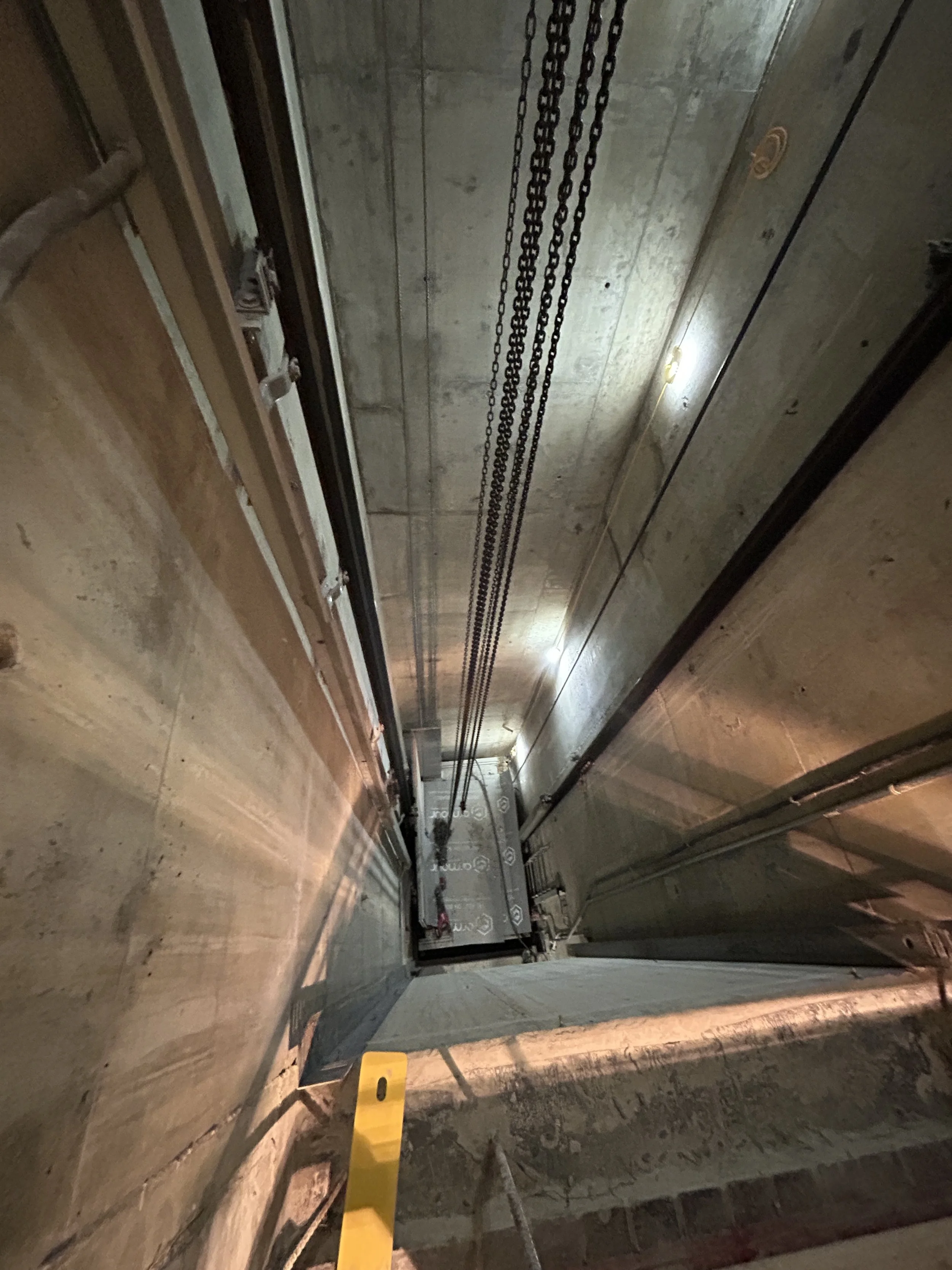 View down a concrete elevator hoistway with guide rails and chains during construction