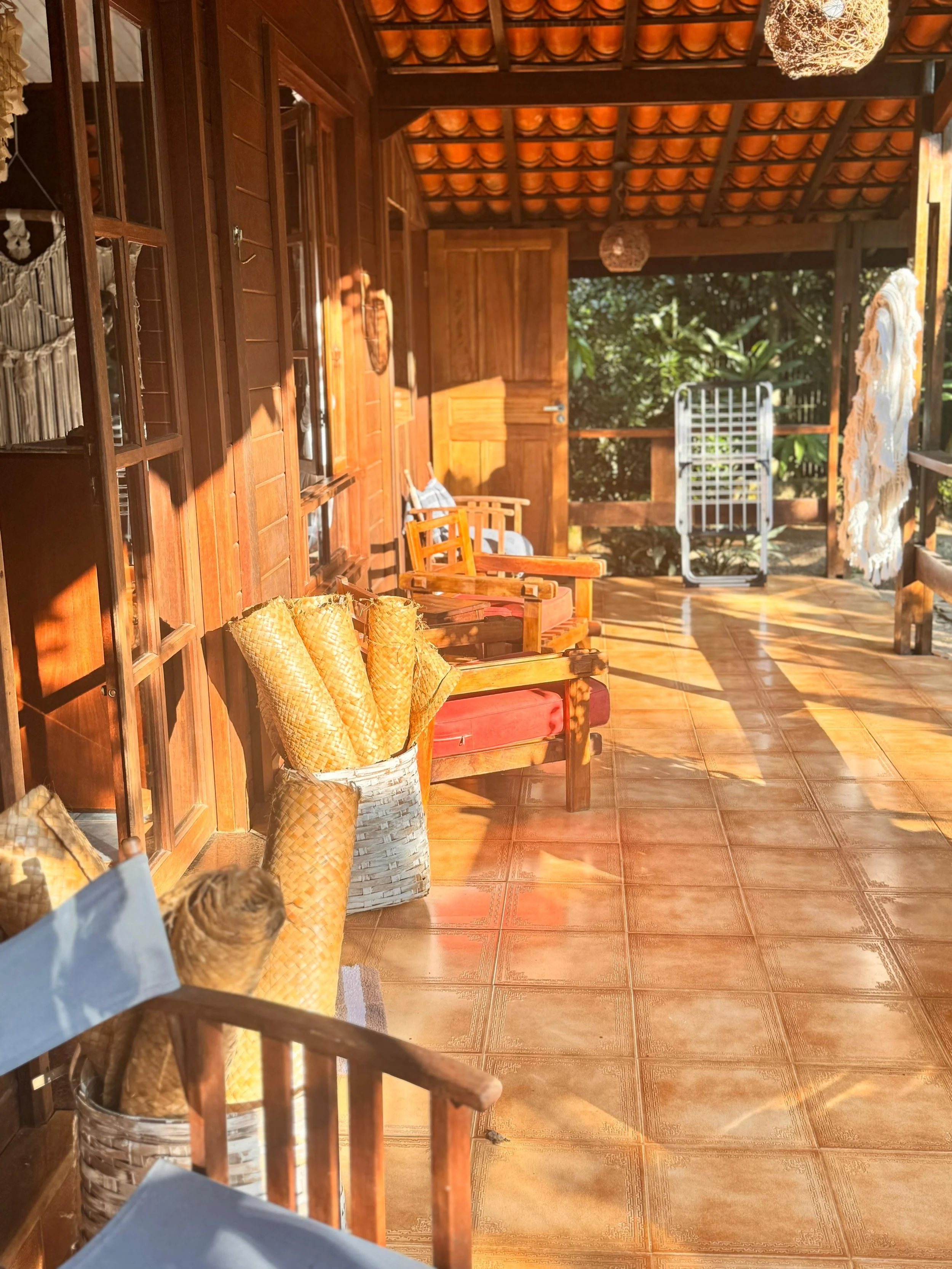 Porche de madera con sillas de madera, cojines, cestas con rollos de papel craft, decoraciones y un área con vista a la naturaleza, iluminado por luz solar.