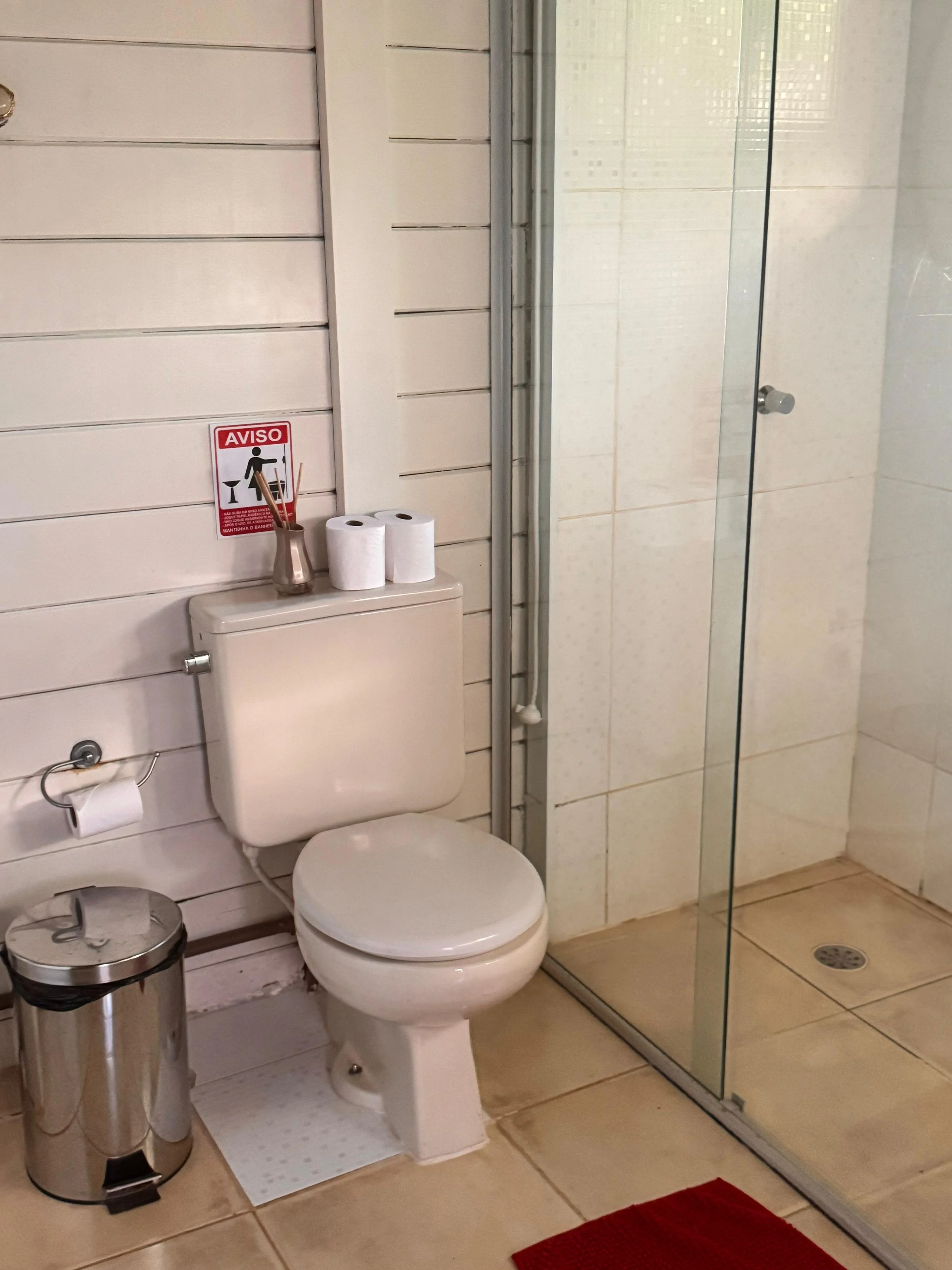 Baño con inodoro, papel higiénico, basura, ducha con puerta de vidrio y pisos de cerámica.