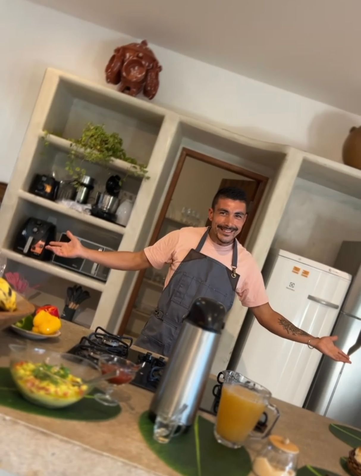 Hombre en la cocina con brazos abiertos, rodeado de alimentos y utensilios, sonriente.