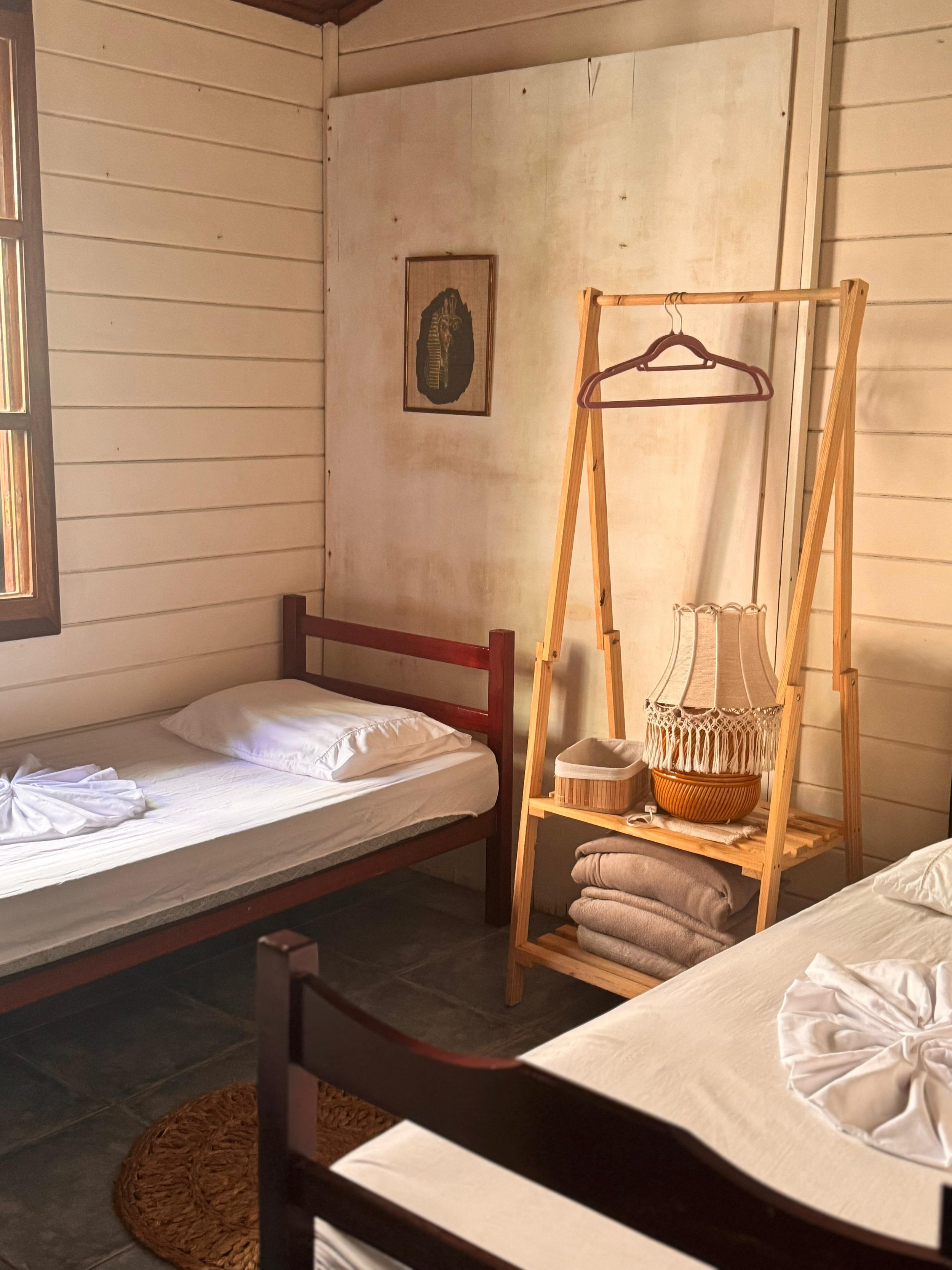 Dormitorio con dos camas simples, paredes de madera pintadas de blanco, una ventana, una lámpara de papel con flecos, y una estructura de madera para colgar ropa con varias toallas dobladas debajo.