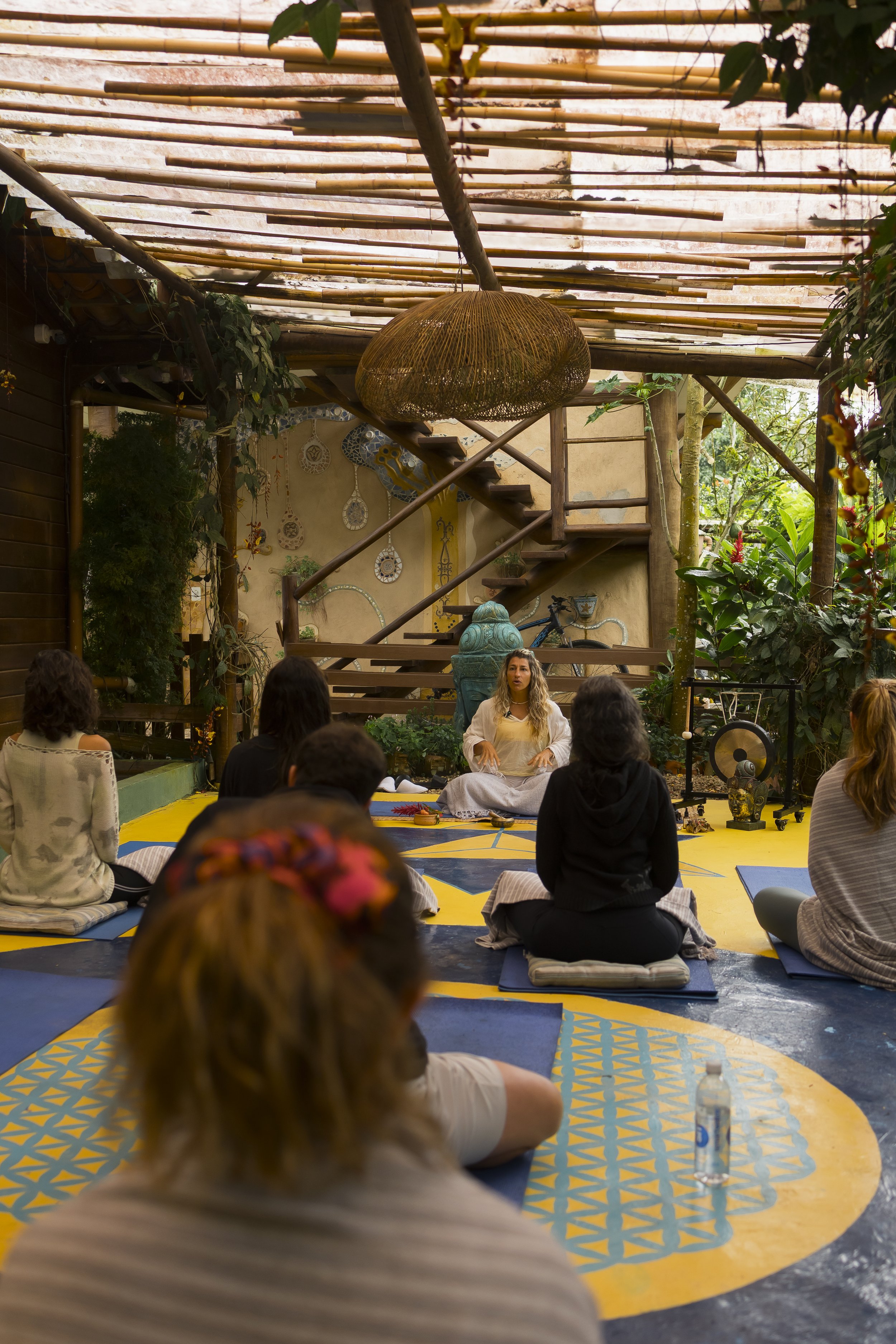 Grupo de personas participando en una clase de meditación o yoga en un espacio abierto con plantas y decoraciones artesanales.