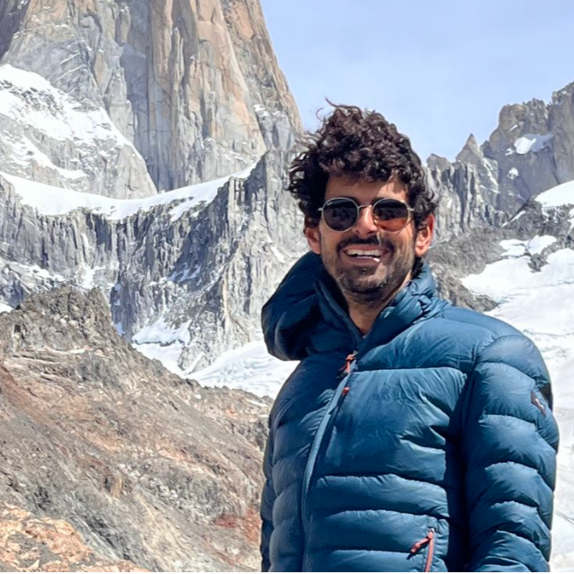 Hombre sonriendo con gafas de sol, lleva un abrigo azul y está en un entorno de montañas con nieve y rocas grandes.