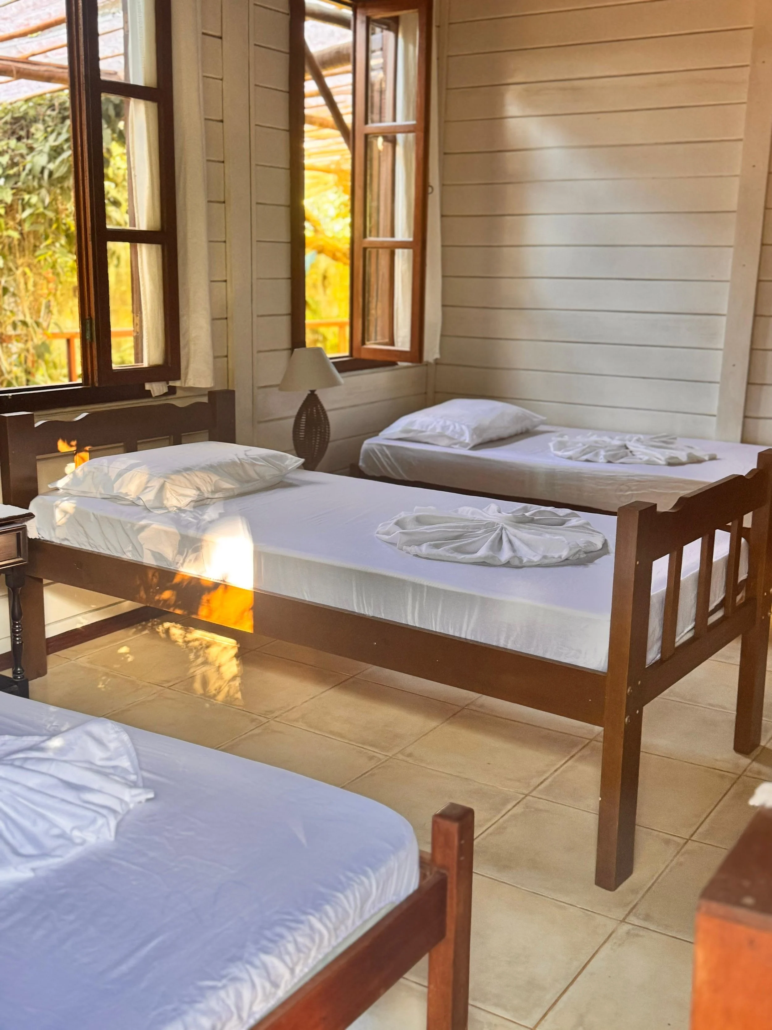 Habitación con paredes de madera, con tres camas de madera, ropa de cama blanca, una lámpara en una mesita y ventanas abiertas que dejan entrar luz natural y vistas verdes afuera.