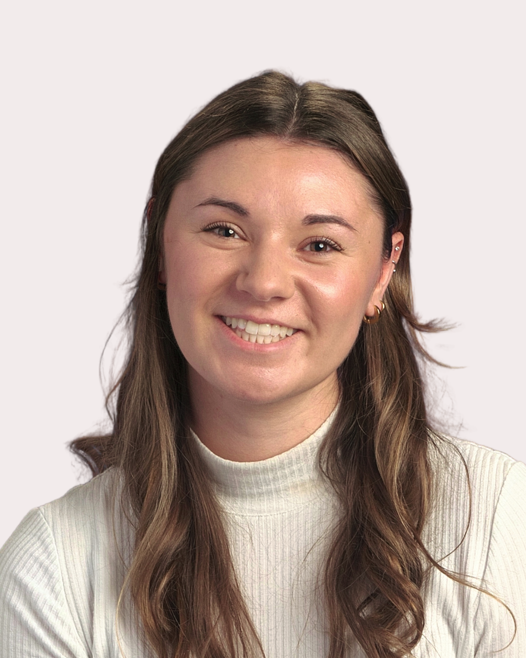 A young woman with long brown hair, smiling, wearing a white high-neck ribbed shirt, earrings, and multiple earrings in her ear, against a plain white background.