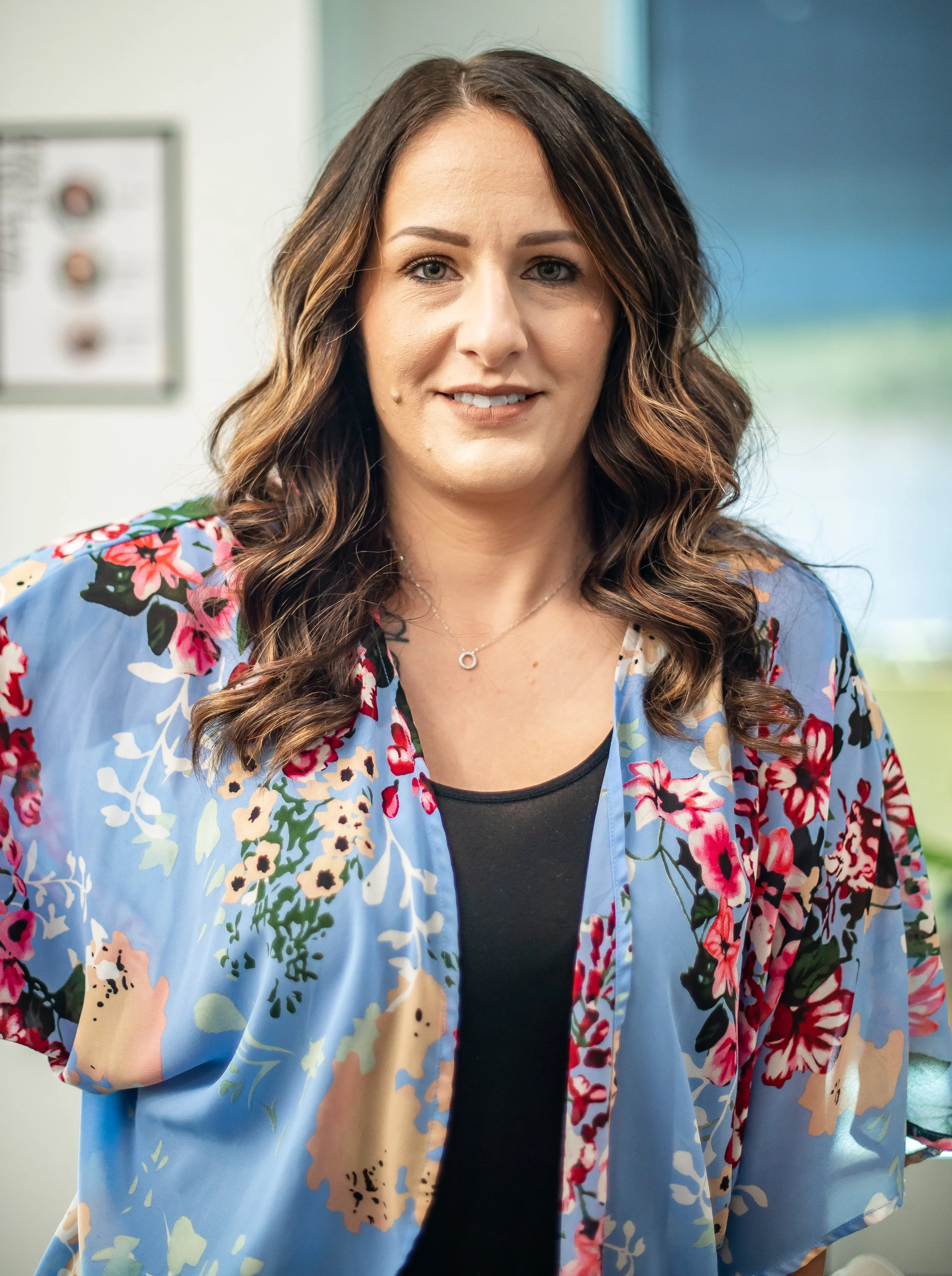 A woman with wavy brown hair wearing a floral blue and pink cardigan over a black top, standing indoors with a window in the background.