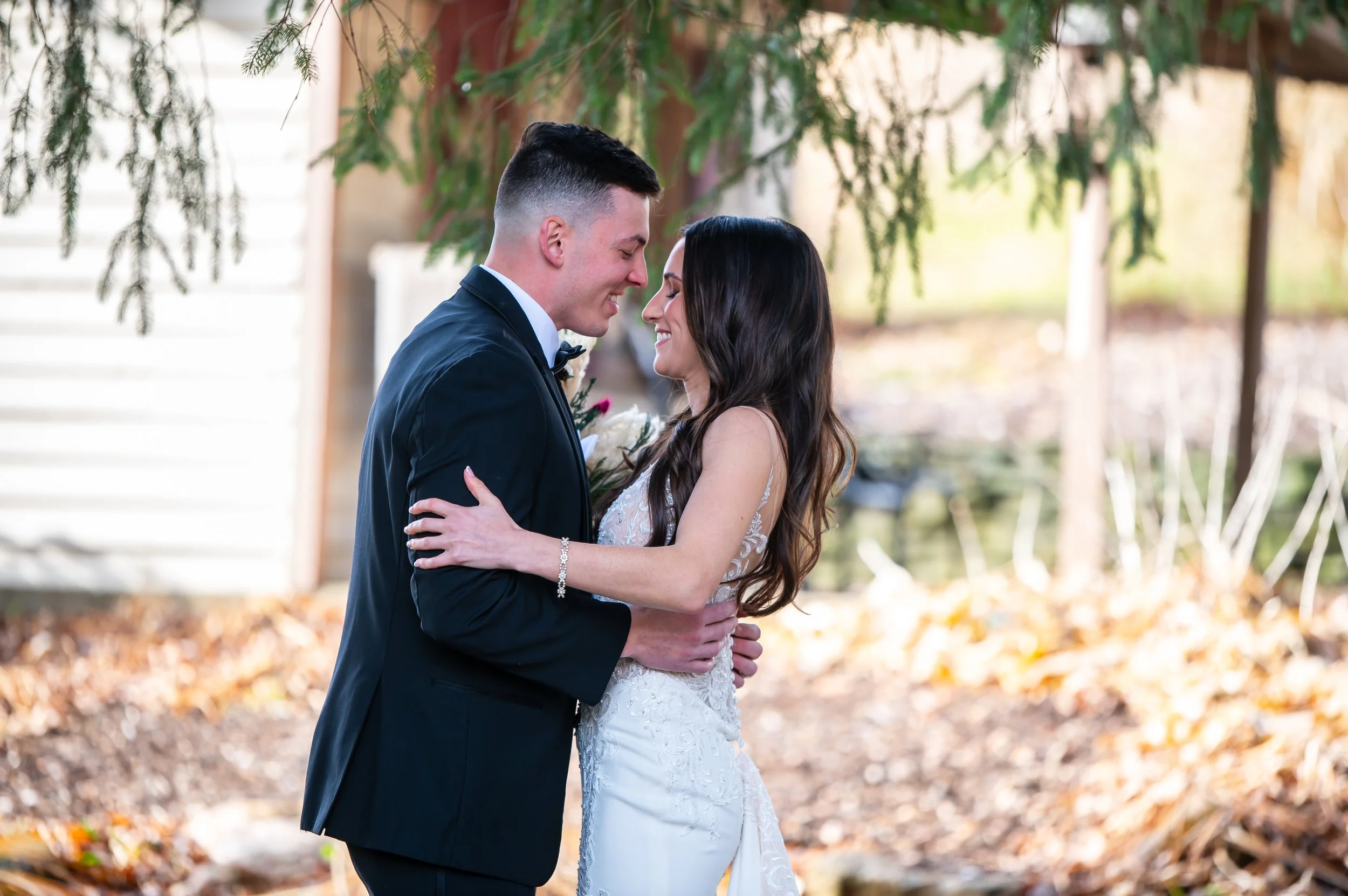 A bride and groom hold each other close and smile during their outdoor wedding, with trees and fallen leaves in the background.