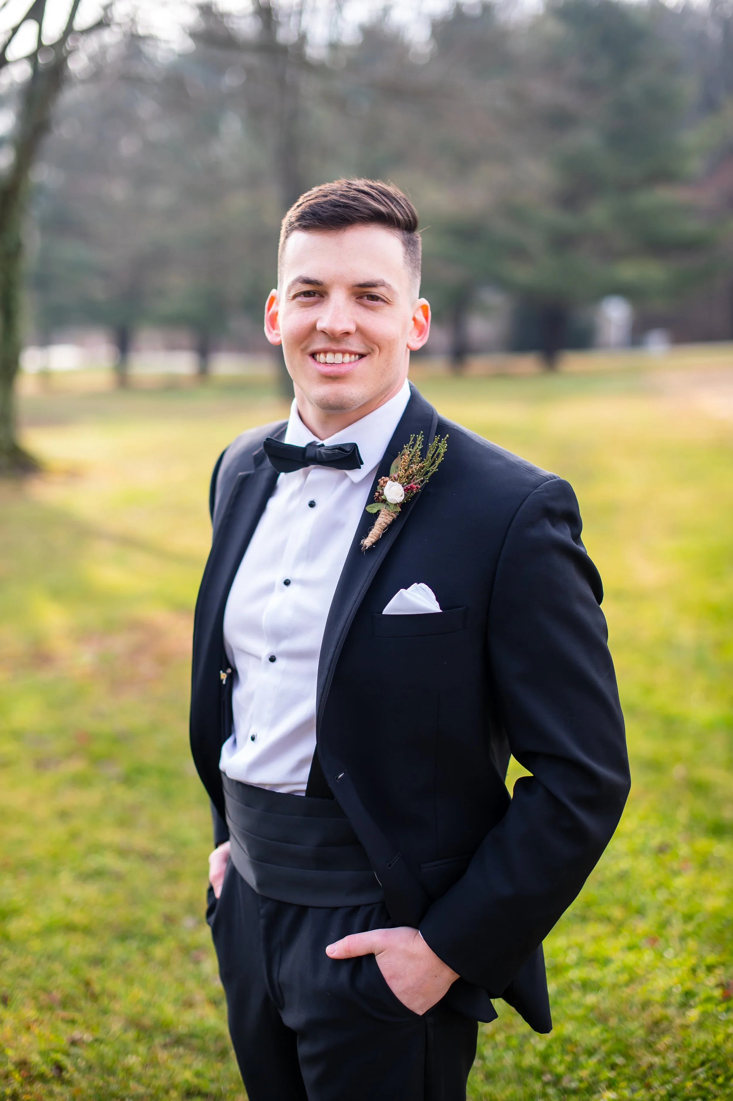 A groom wearing a black tuxedo with a white shirt and black bow tie, standing outdoors on a grassy area with trees in the background, smiling at the camera.