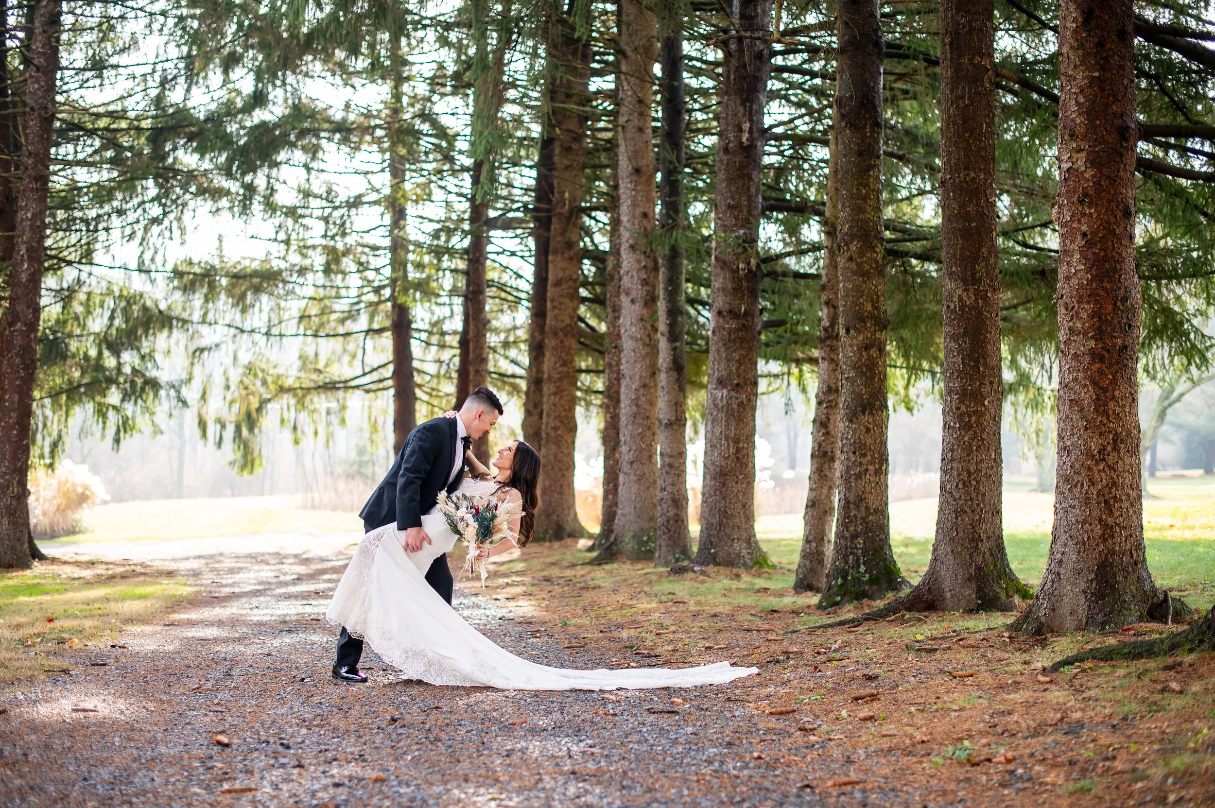 Groom dipping bride while holding her waist, bride holding bouquet, in a wooded outdoor setting with tall pine trees.