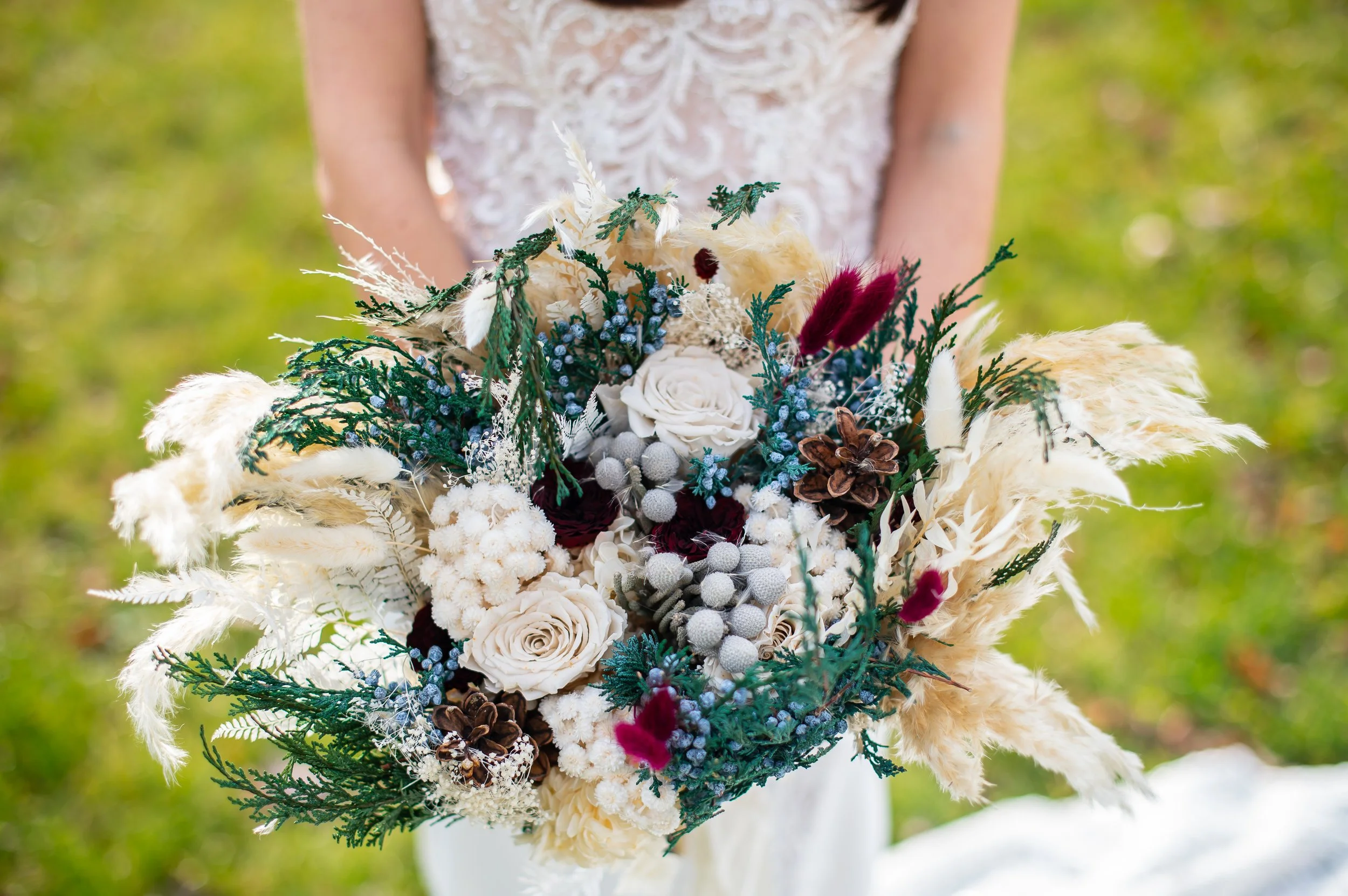 A person in a white lace dress holding a large bouquet of white, burgundy, and green flowers and foliage outdoors.