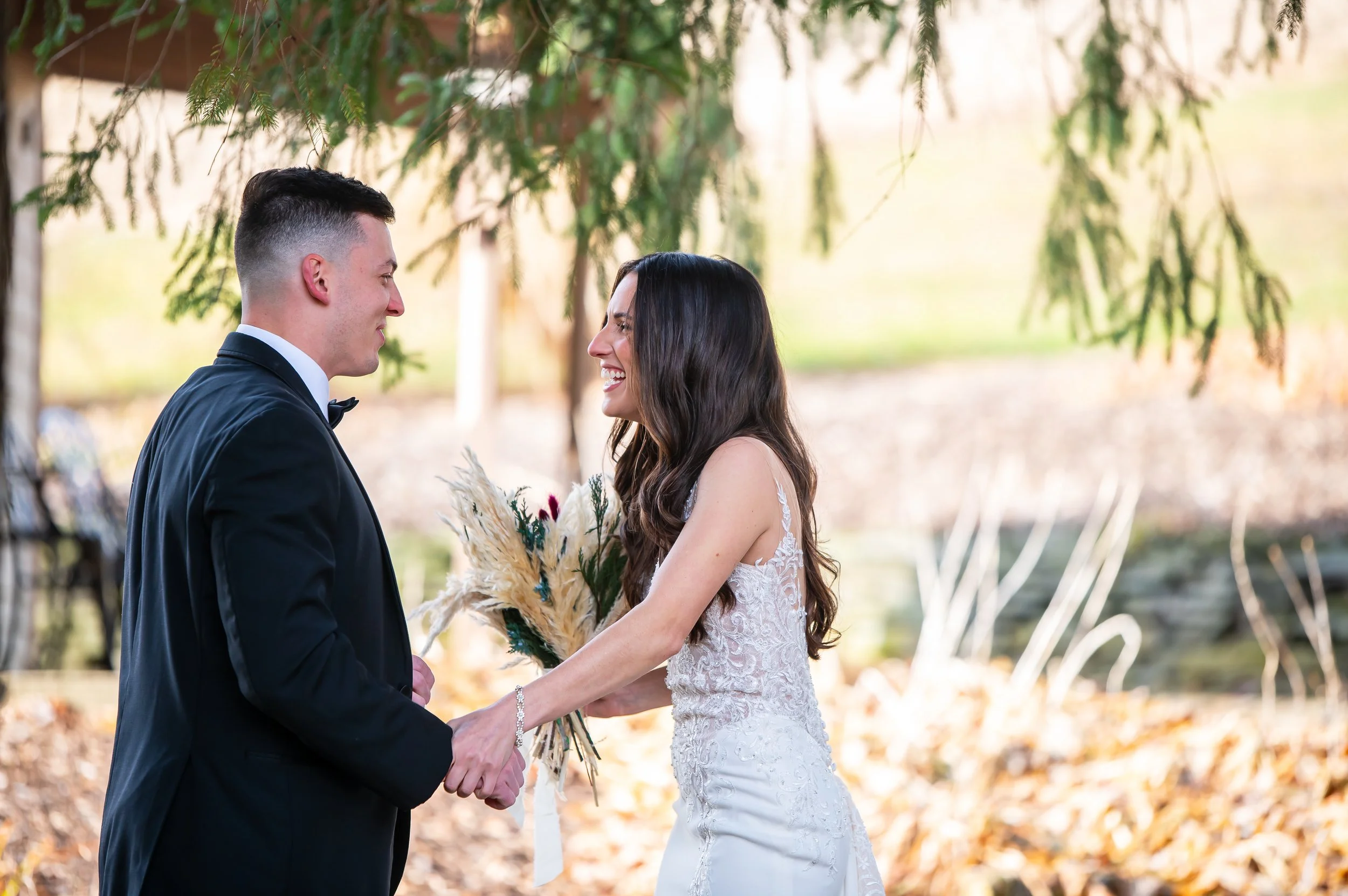 A bride and groom holding hands and smiling at each other outdoors during their wedding, surrounded by trees with fall leaves.