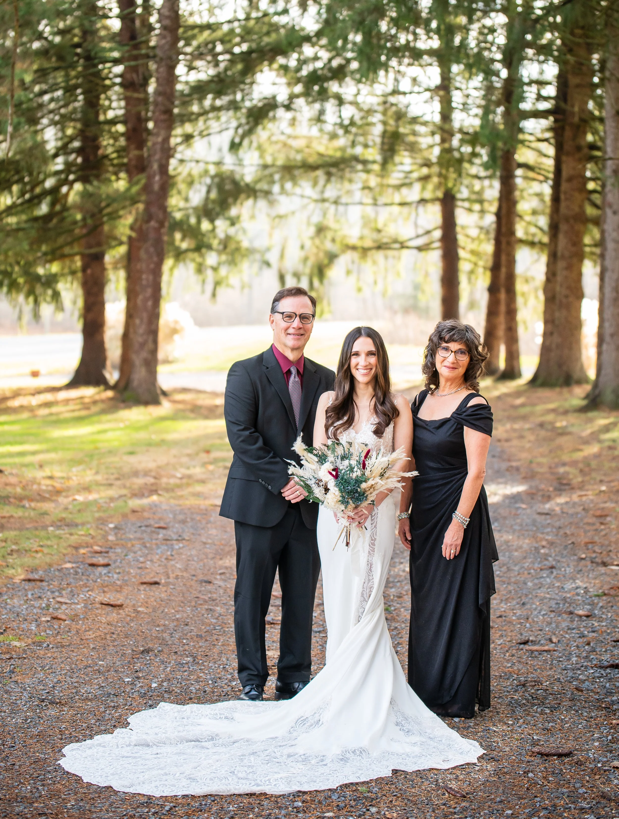 A bride in a white wedding dress holding a bouquet of flowers, standing with her parents in a wooded outdoor setting during daytime.