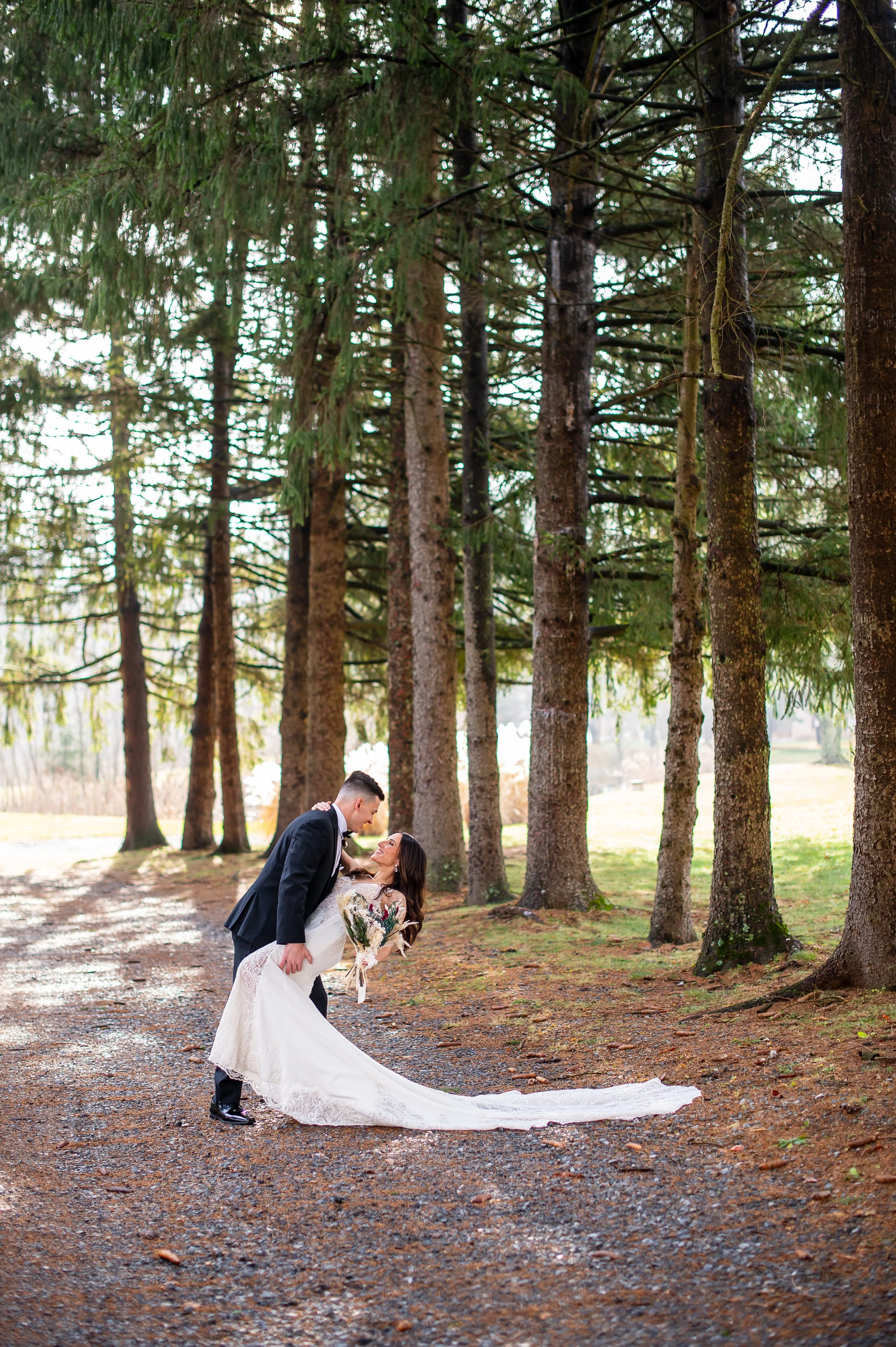 A bride and groom dancing outdoors on a wooded path with tall trees in the background.