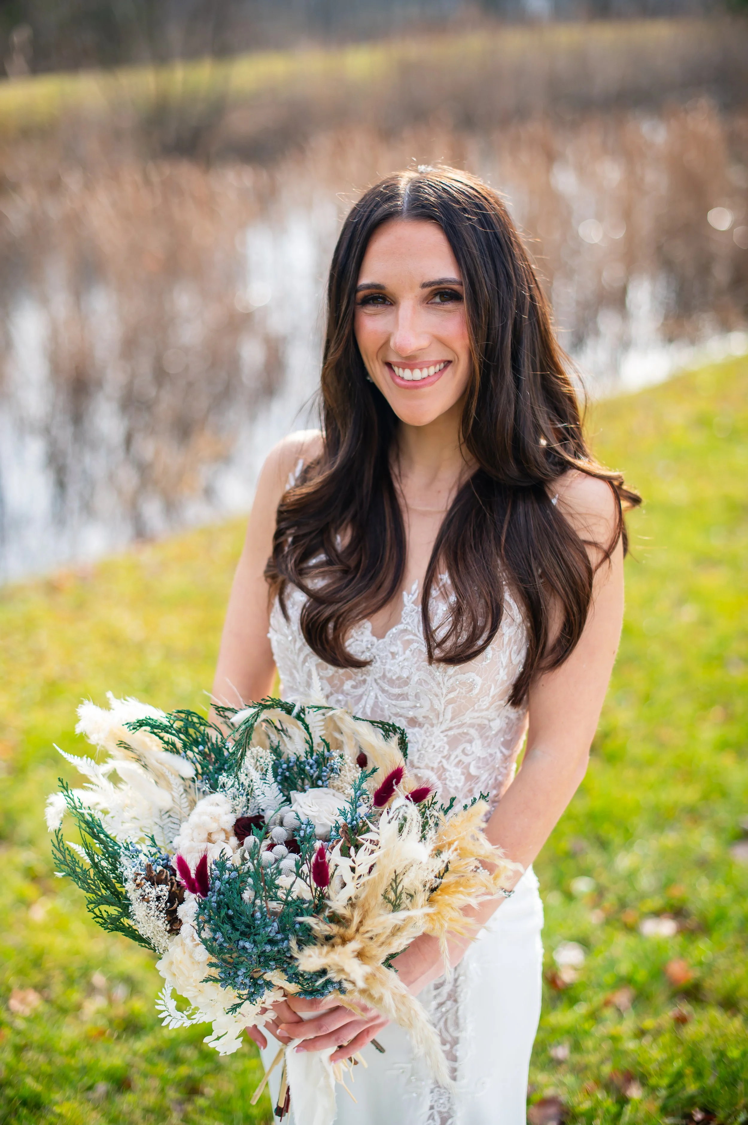 A smiling woman in a white lace wedding dress holding a bouquet of mixed flowers outdoors near a pond with blurred brown reeds and greenery in the background.