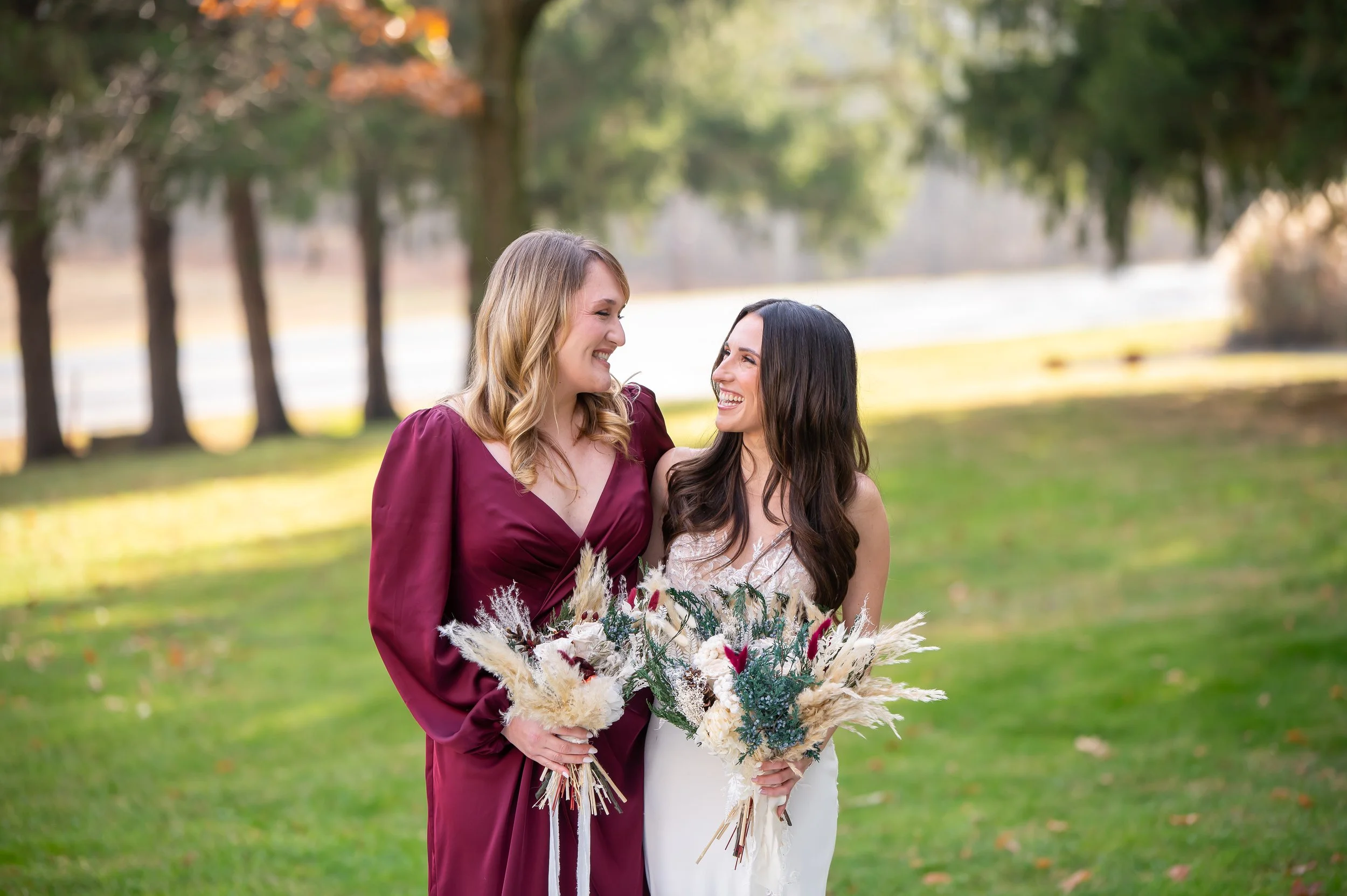 Two women standing outdoors in a park, holding bouquets of dried flowers, smiling and looking at each other. One woman wears a maroon dress, and the other wears a white dress. Trees and grass are in the background.
