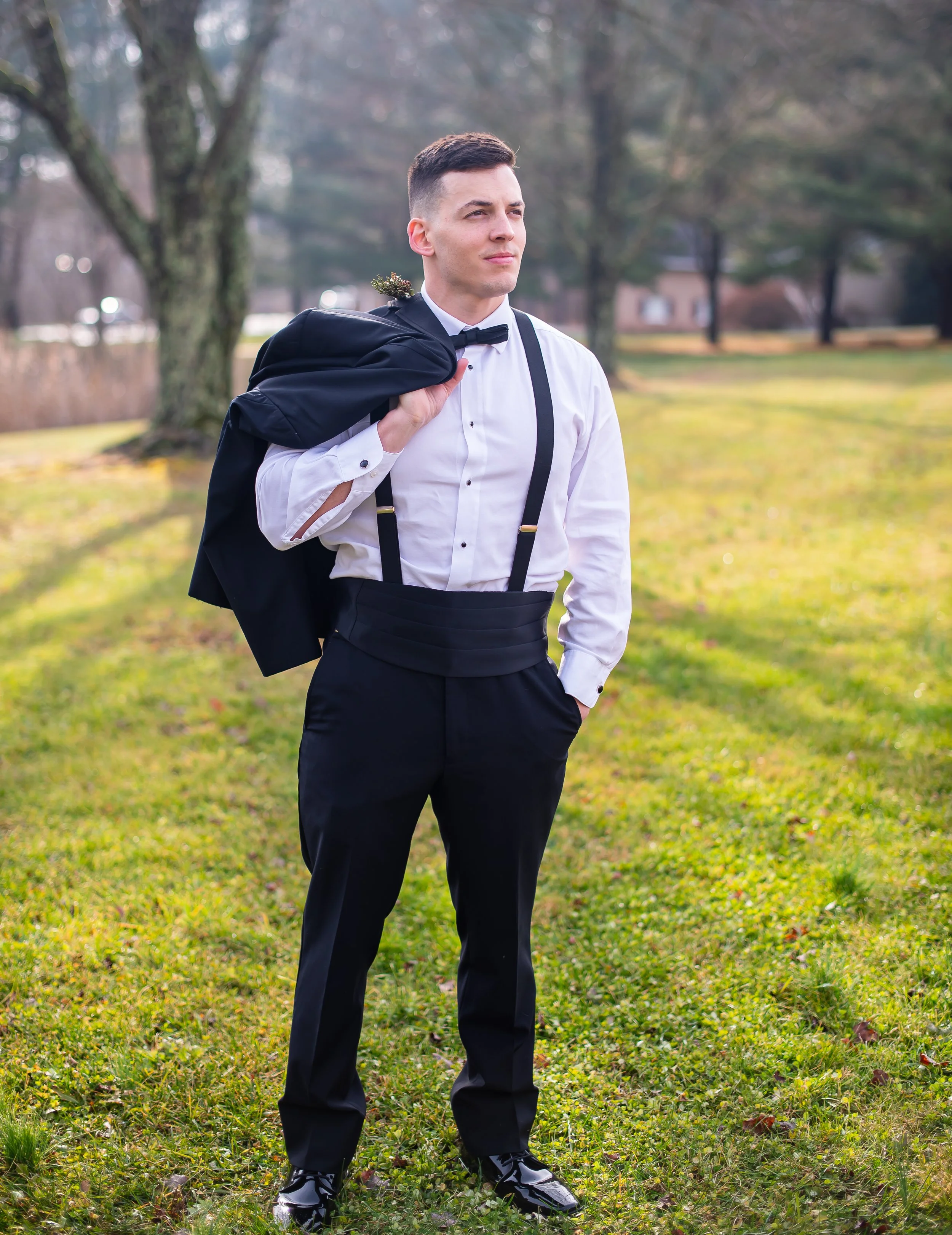 Young man dressed in formal tuxedo with suspenders, carrying a suit jacket over his shoulder, standing outdoors on grass with trees and houses in the background.