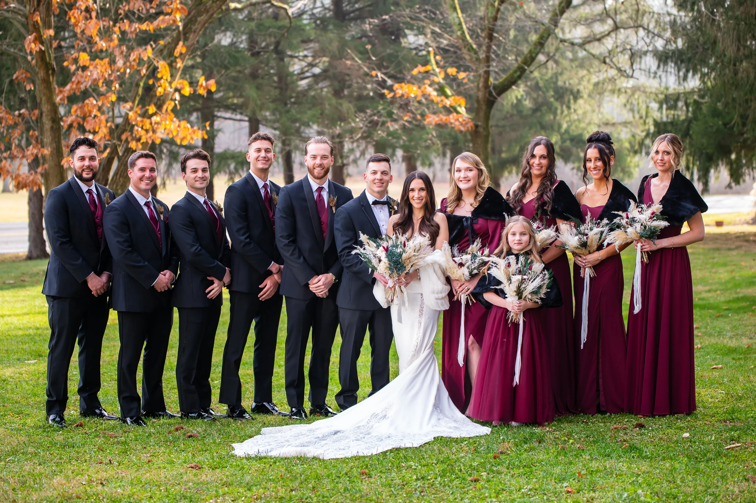 A wedding party standing outdoors on grass, including the bride in a white dress, groom in a black tuxedo, bridesmaids in maroon dresses, and groomsmen in dark suits, all holding bouquets or boutonnieres, with trees and autumn leaves in the backgroun