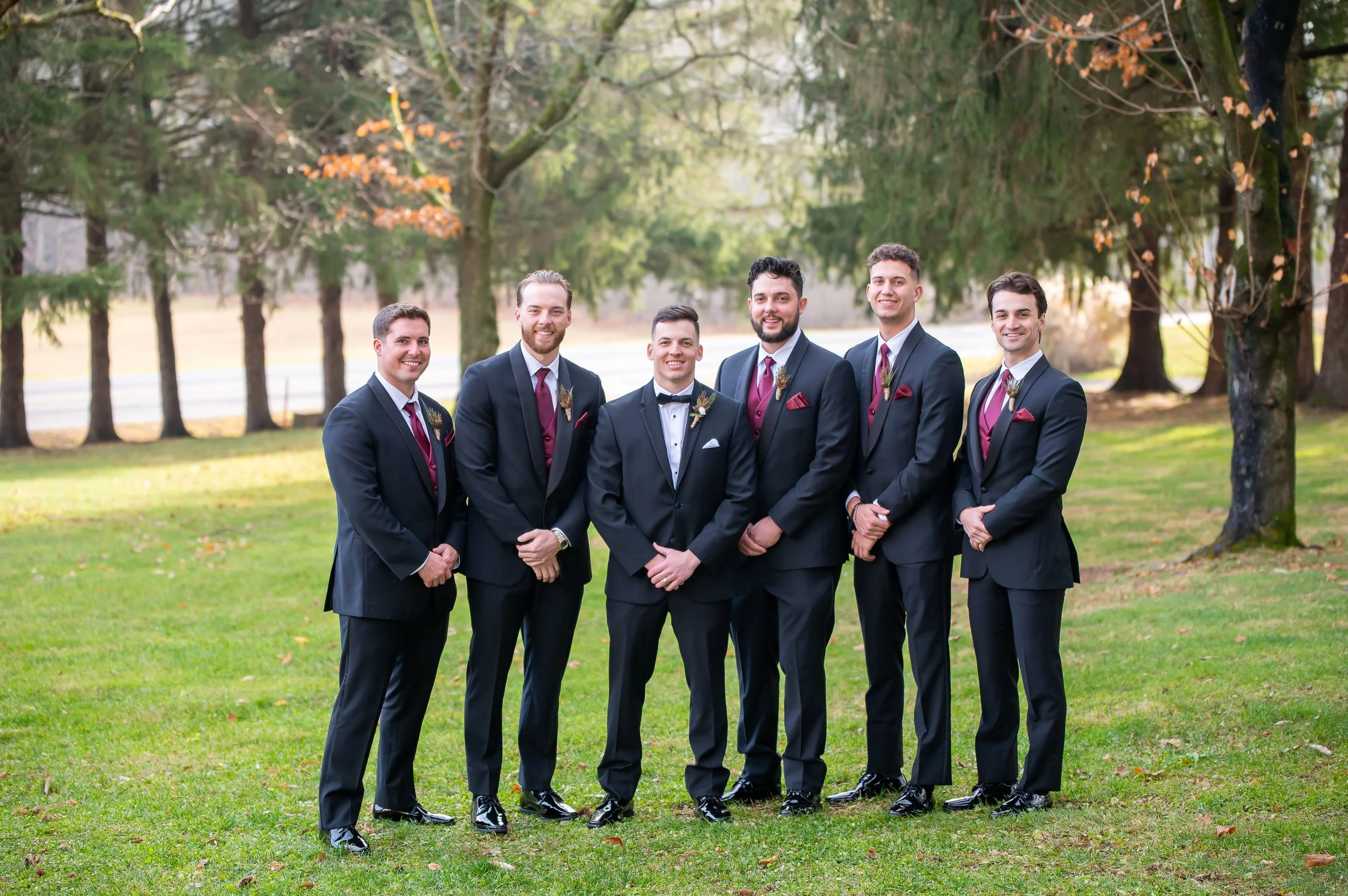 Group of seven men dressed in suits standing outdoors on a grassy area with trees in the background, smiling for a photo.