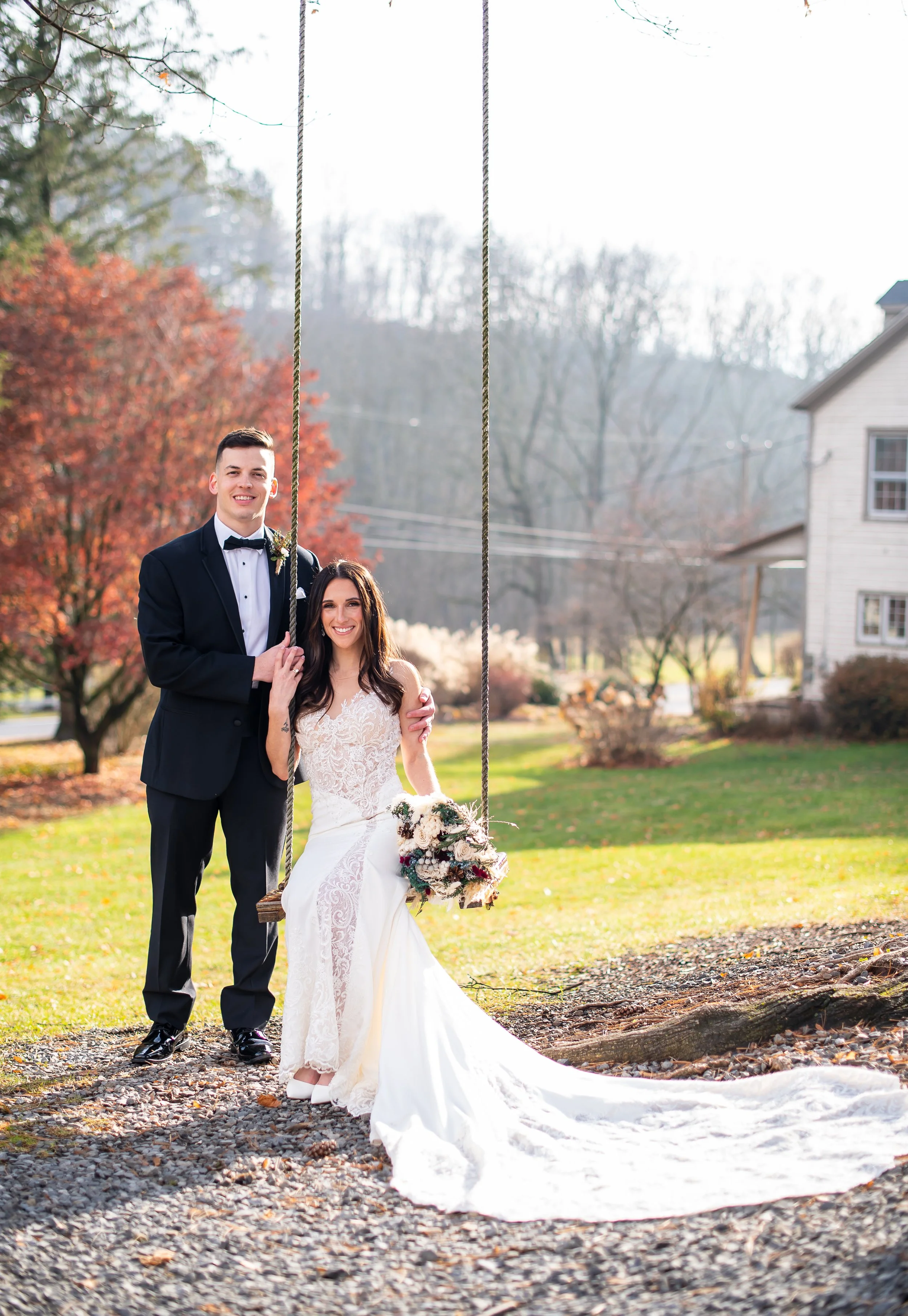A bride and groom pose outdoors near a swing, with autumn trees and a house in the background. The groom wears a black tuxedo, and the bride wears a white lace wedding gown holding a bouquet.